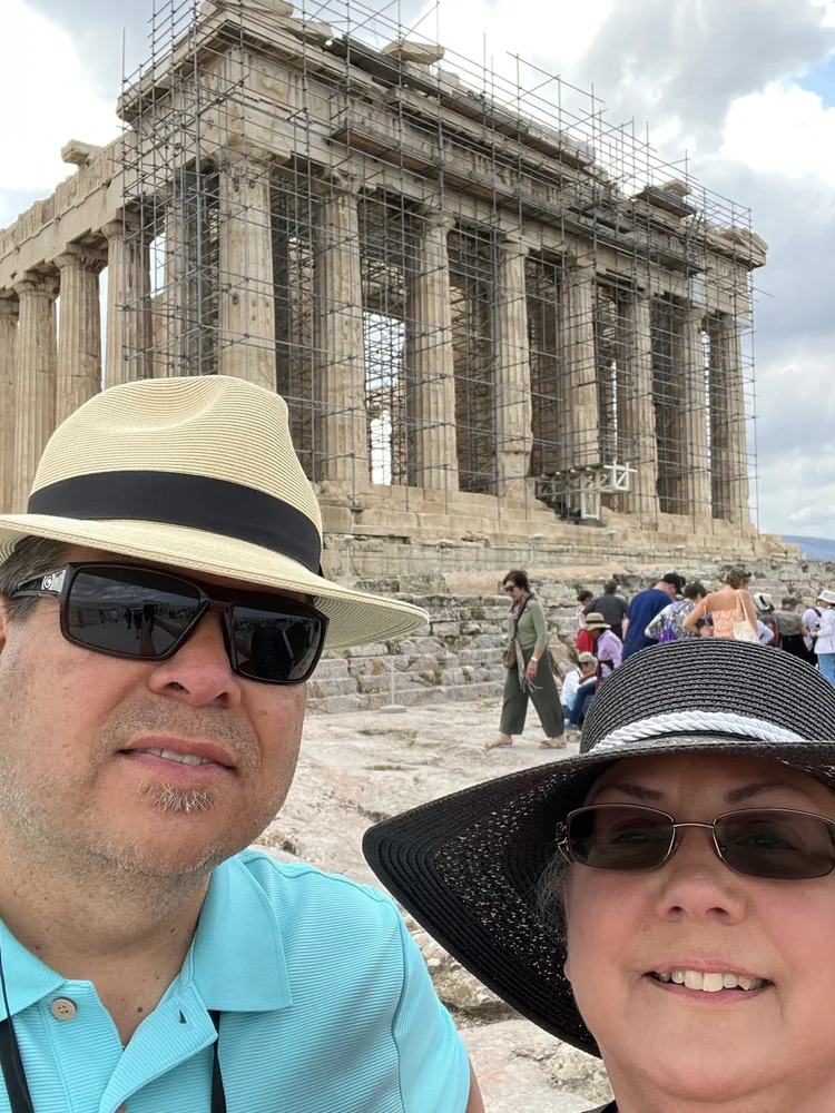 A middle-aged man and woman wearing sunglasses and sun hats taking a selfie in front of the Parthenon on the Acropolis in Athens, Greece. The Parthenon is under renovation with scaffolding around it, and there are other tourists in the background.