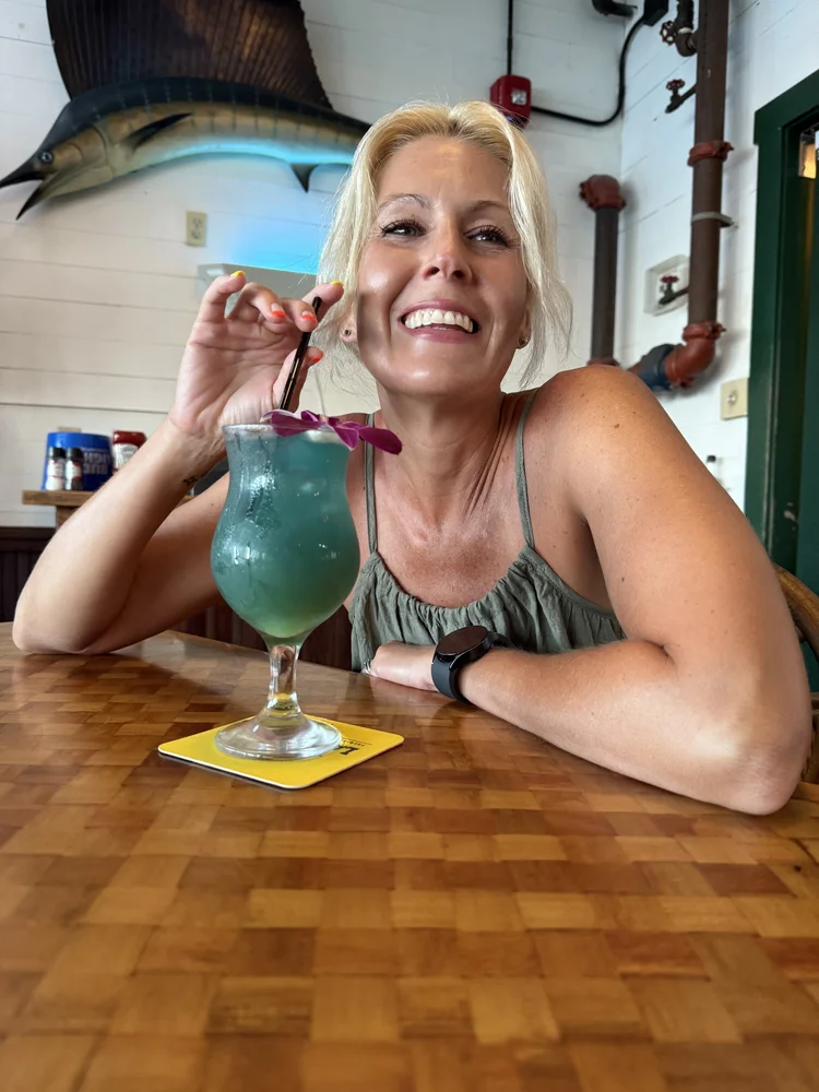 A woman with blonde hair smiling at the camera while holding a blue cocktail with a purple flower garnish in a restaurant or bar setting, with a large fish mounted on the wall behind her.
