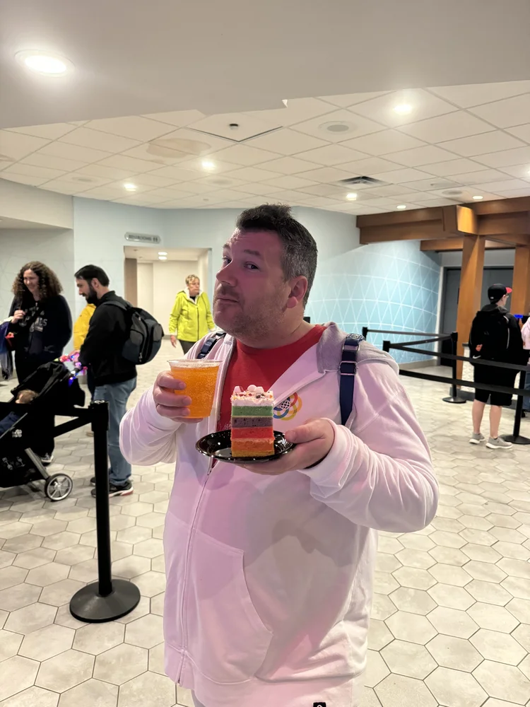 A man holding a plate with a slice of layered rainbow cake and a cup of orange beverage in an indoor setting with other people in the background.
