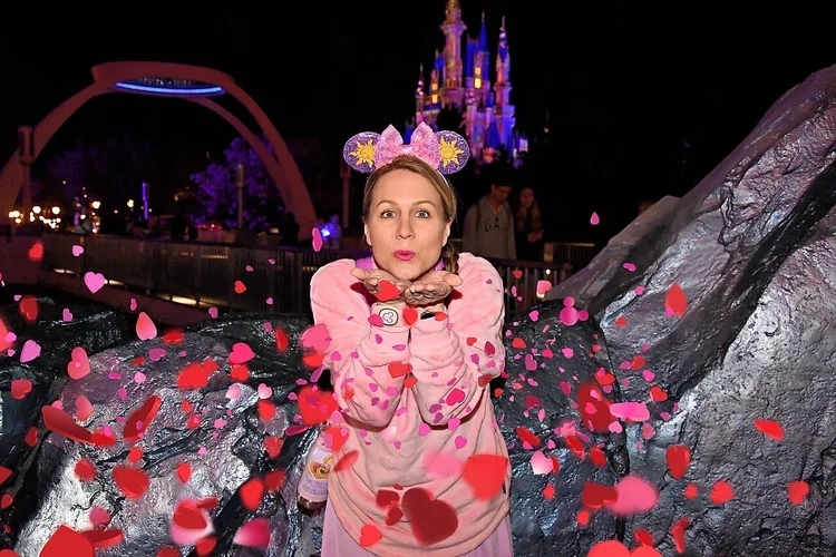 A woman blowing kisses with heart-shaped confetti falling around her at a theme park at night, with a castle illuminated in the background.