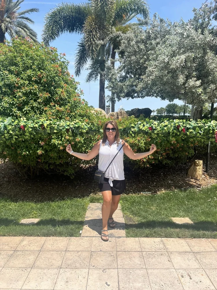 A woman smiling and posing on a stone path in front of lush green bushes and palm trees on a sunny day.