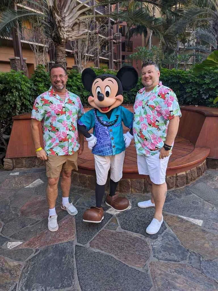 Two men in colorful Hawaiian shirts standing next to a person dressed as Mickey Mouse in a tropical setting with palm trees and greenery.