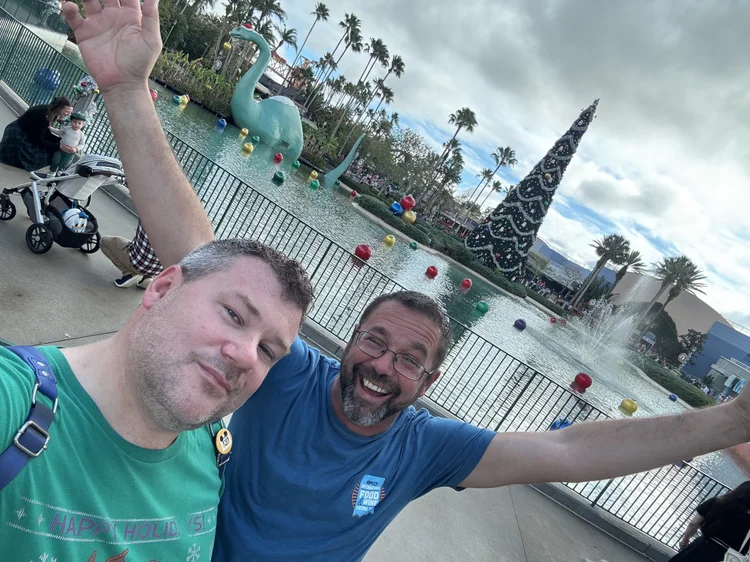 Two men taking a selfie in front of a pond with a large holiday-themed Christmas tree, a giant swan sculpture, and colorful floating balls.
