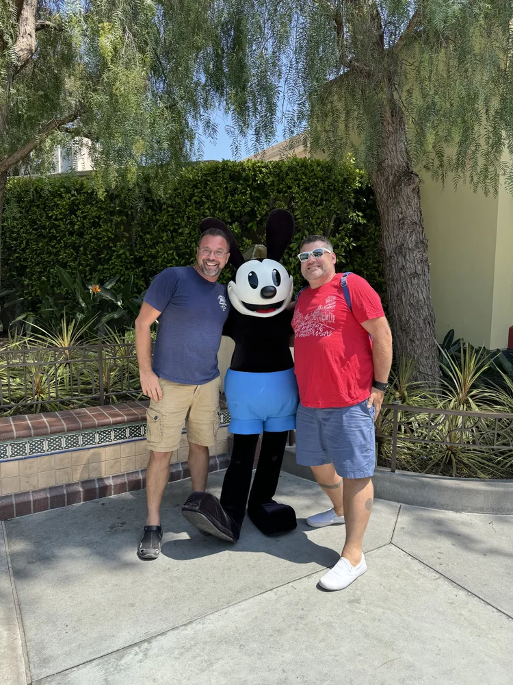 Two men posing with Mickey Mouse mascot outdoors on a sunny day, with trees and bushes in the background.
