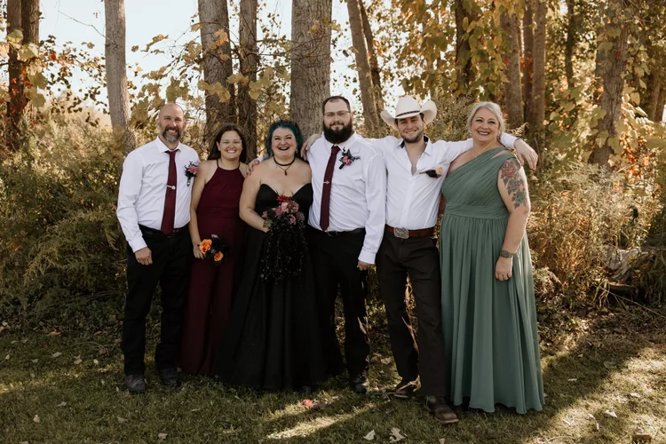 Group of seven people standing outdoors in front of trees, dressed in formal and semi-formal attire for a wedding celebration, with sunlit autumn foliage in the background.