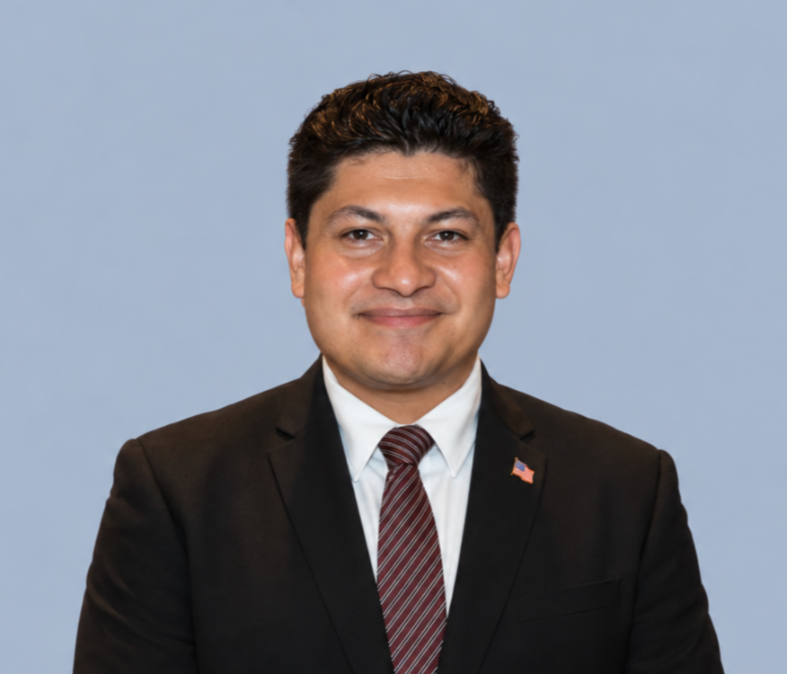 Professional headshot of a man in a black suit, striped tie, and American flag lapel pin against a light blue background