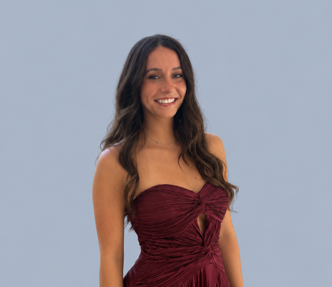 Smiling woman with long wavy dark hair wearing a burgundy ruched dress against a light blue background