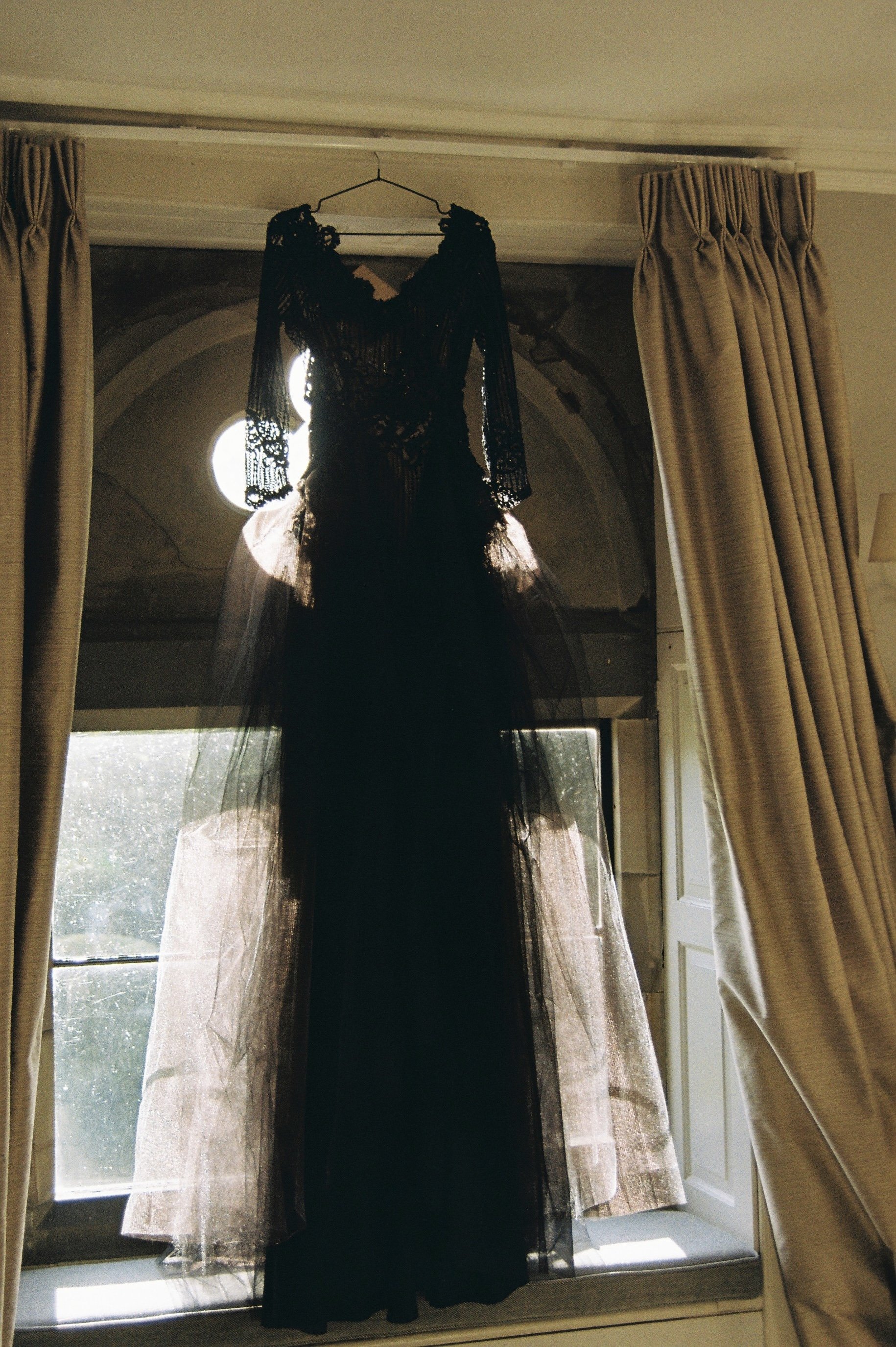 A black lace dress hanging on a hanger in front of a window with curtains, backlit by sunlight.