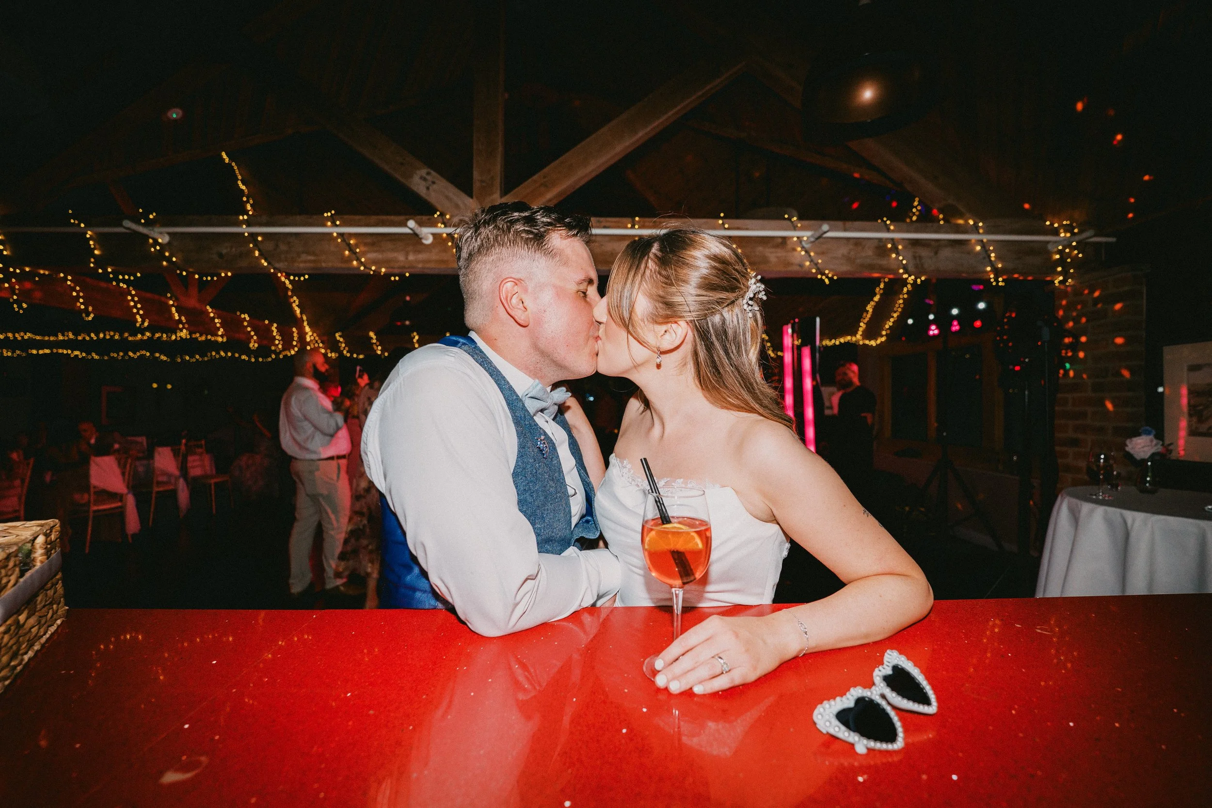 Bride and Groom stood at red bar sharing a kiss. Party and fairy lights are in the background. Venue is the Dodford Inn