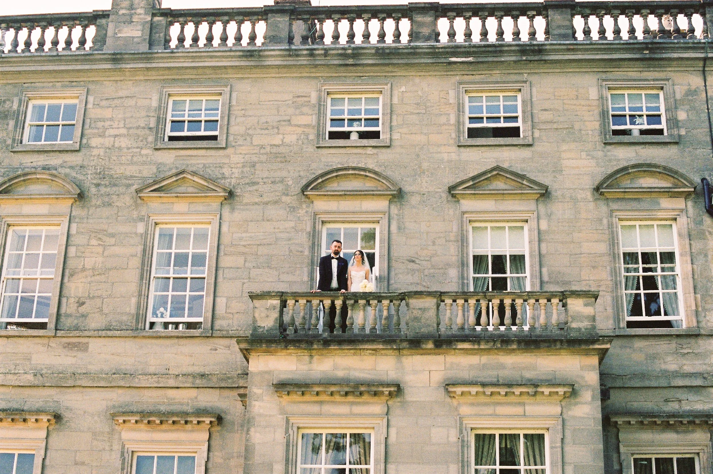 A bride and groom stand on a small balcony outside a historic stone building with multiple windows, during their wedding day.