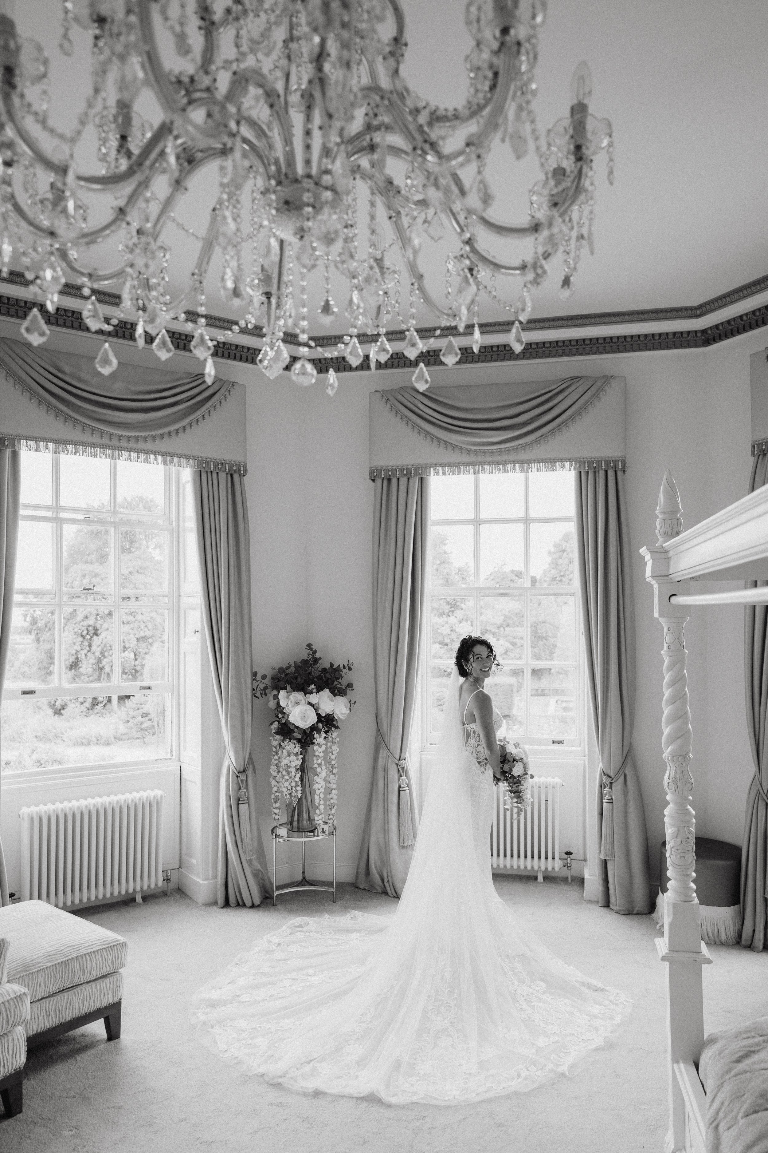 Black and white photo, bride is stood in a large grand room with large windows and a chandelier. At Bourton Hall wedding venue