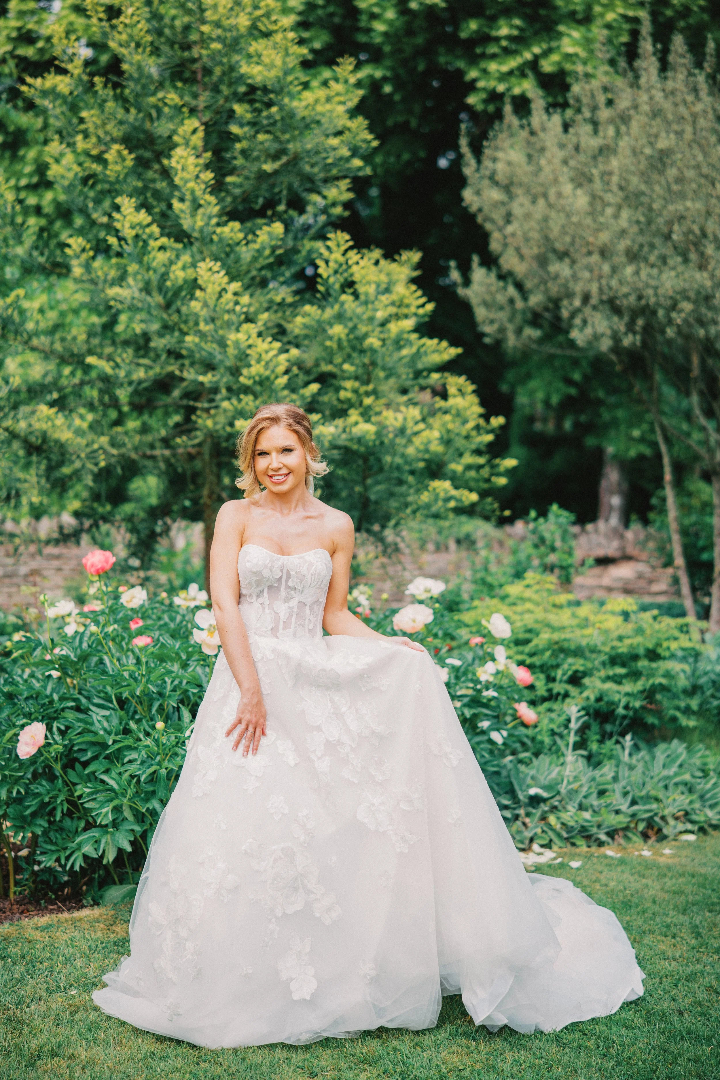 Bride stood on grass surrounded by trees and flowers, the wedding dress has flower details and the woman is smiling, At Birtsmorton Court Wedding venue
