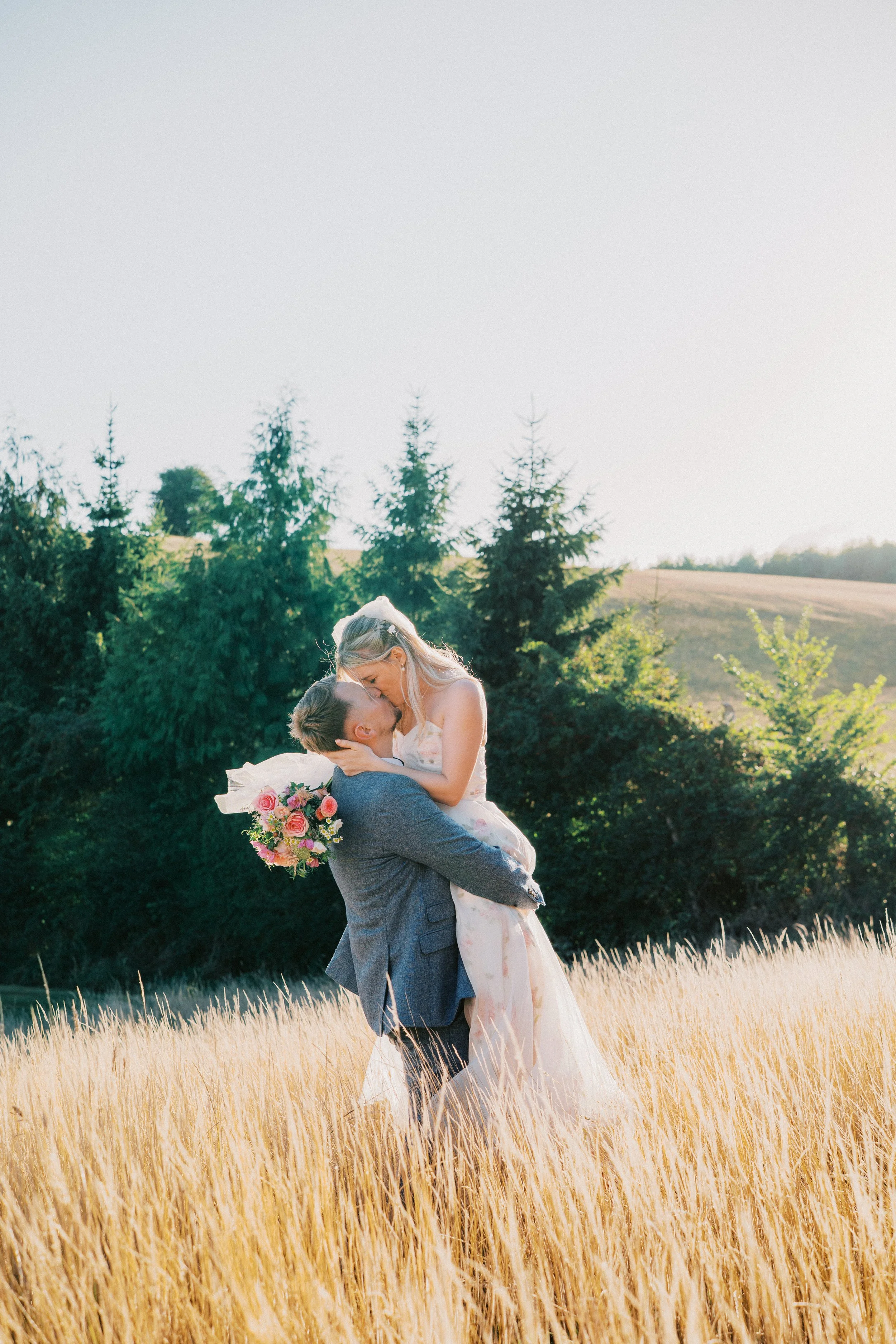 Wedding Couple at Golden Hour sharing a kiss in a field with trees and the sunlight behind them