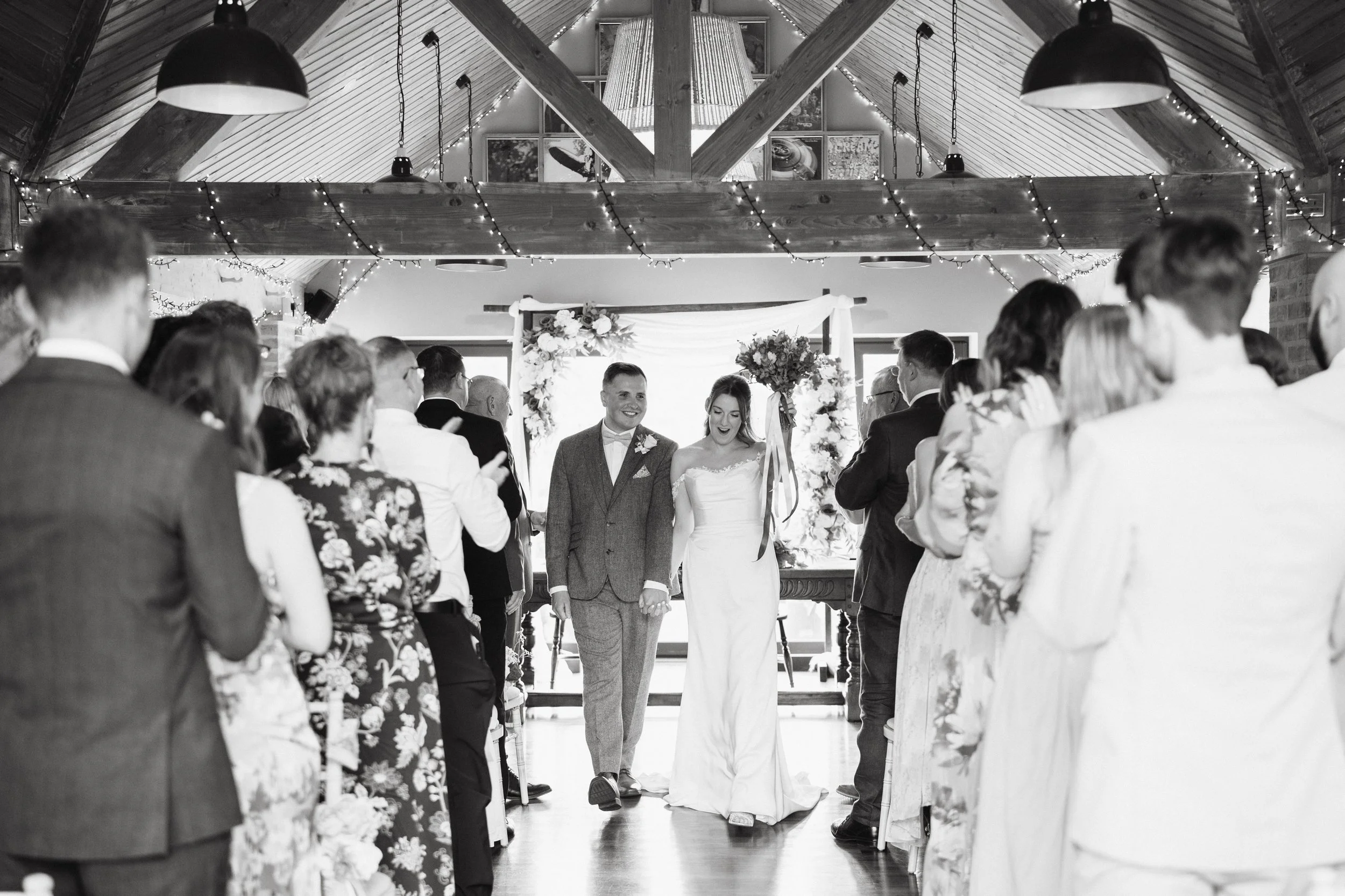 black and white photo of bride and groom walking back down the aisle together cheering after they've just got married in large room with wooden beams and fairy lights