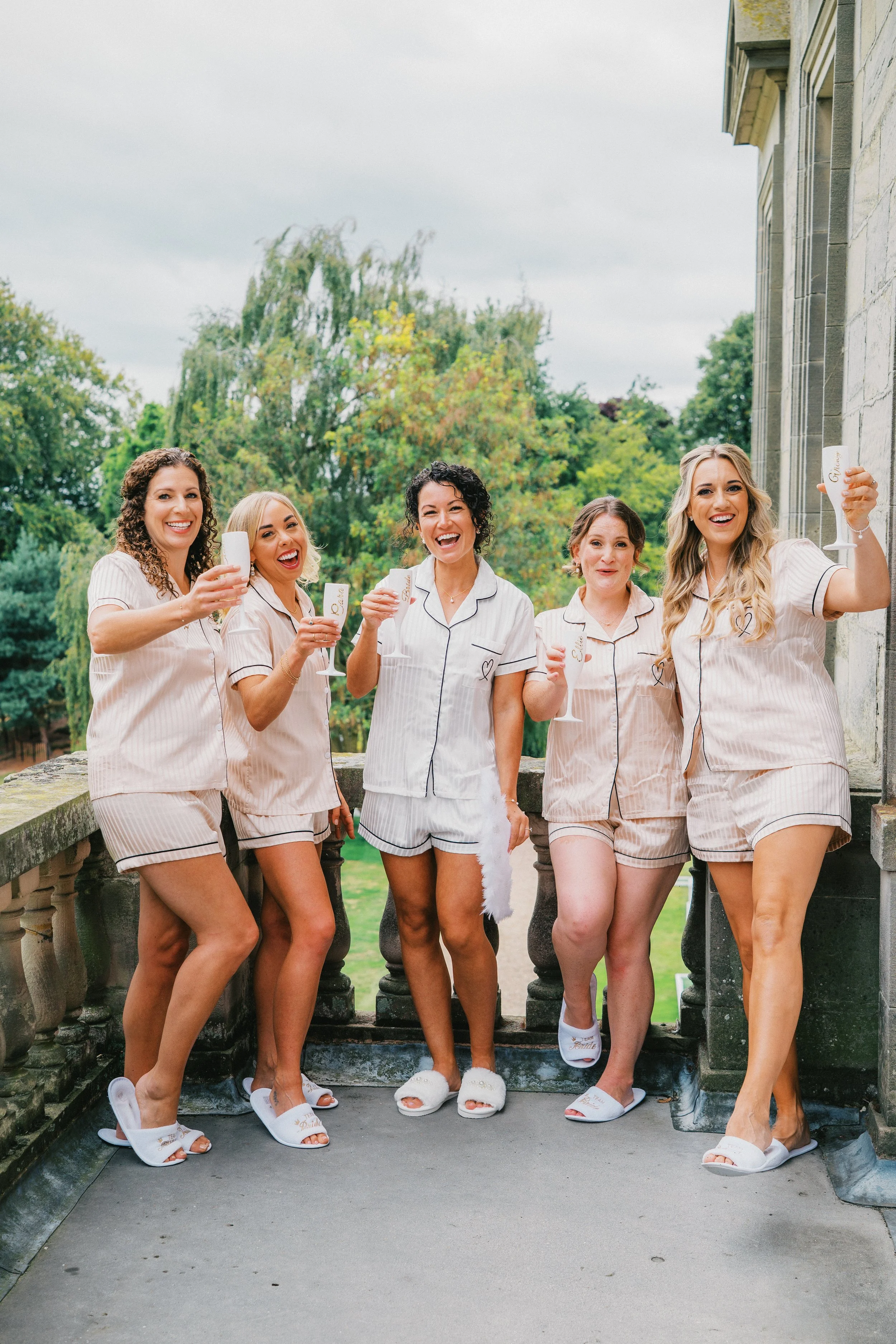 Bride and bridesmaids in pyjamas stood on a balcony cheering their drinks