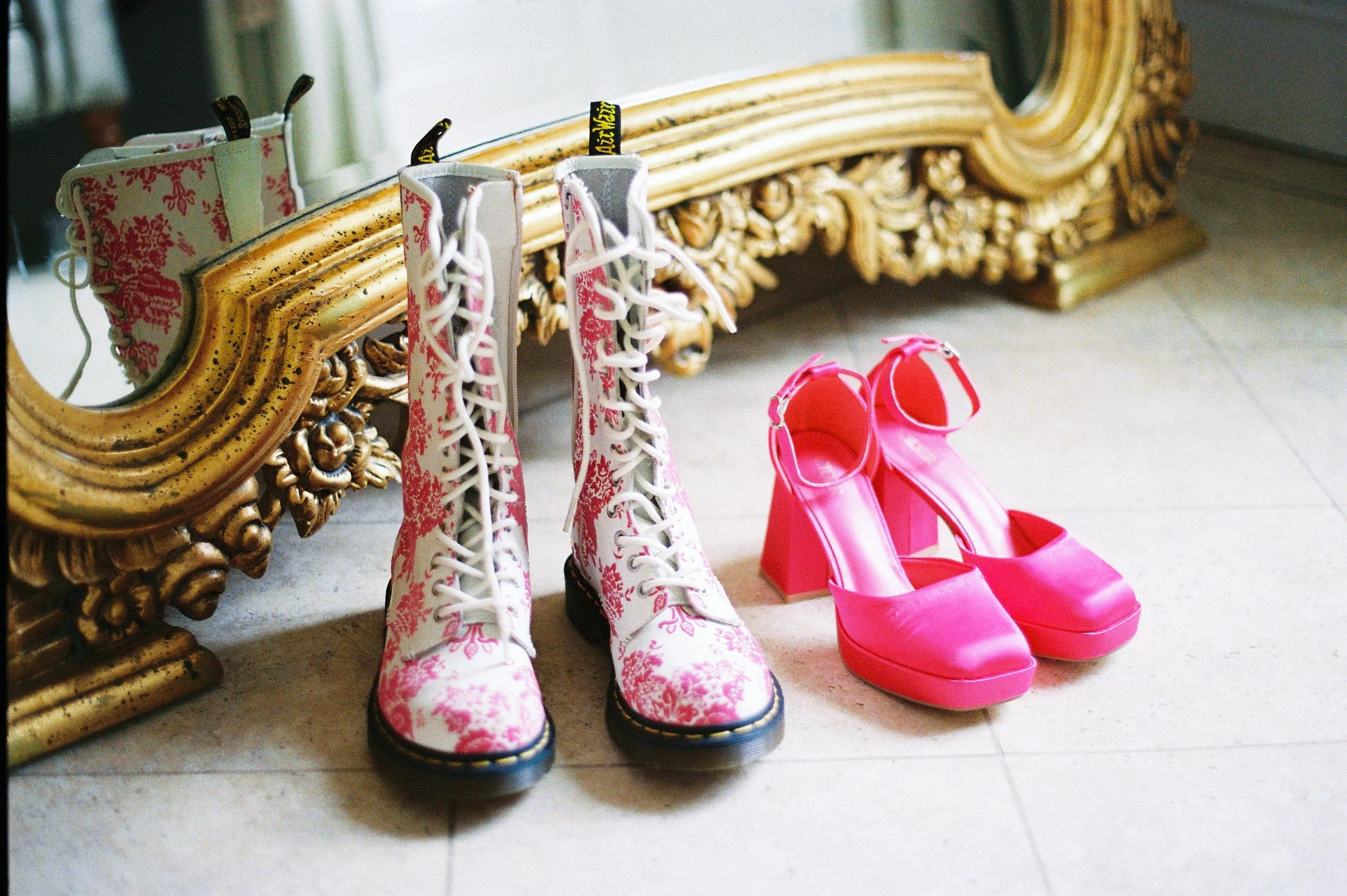 Pair of white boots with pink floral pattern and black soles, and pair of bright pink high heel shoes, placed on a tiled floor against an ornate gold-framed mirror.