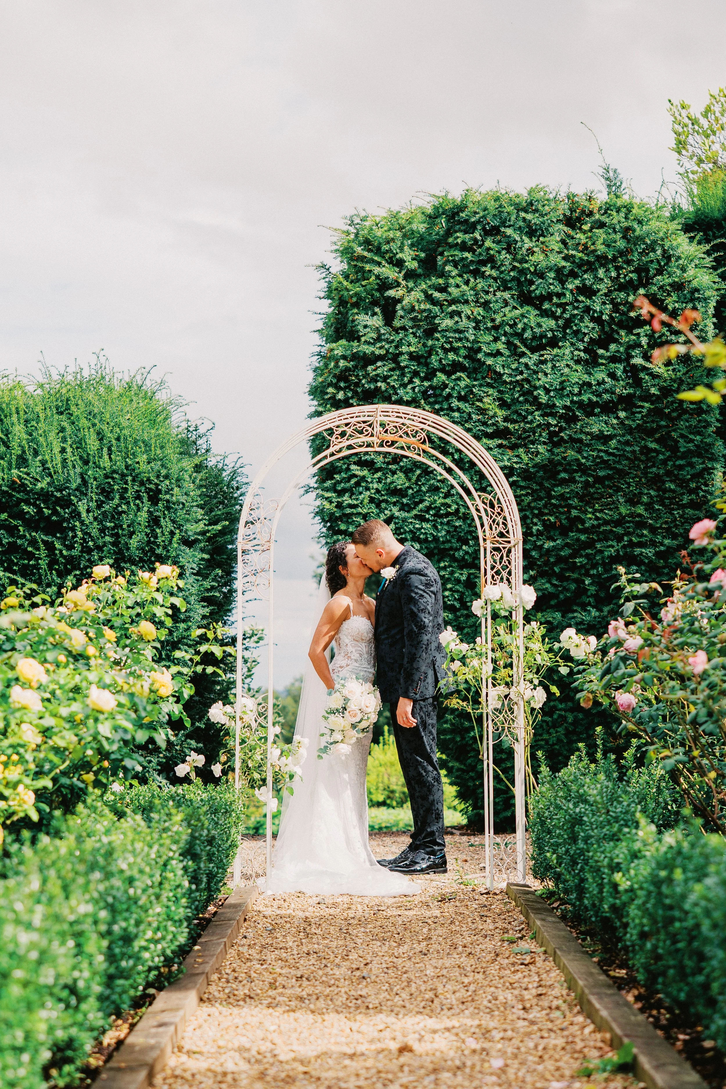 bride and groom kissing under archway surrounded by green bushes and trees and a rose garden