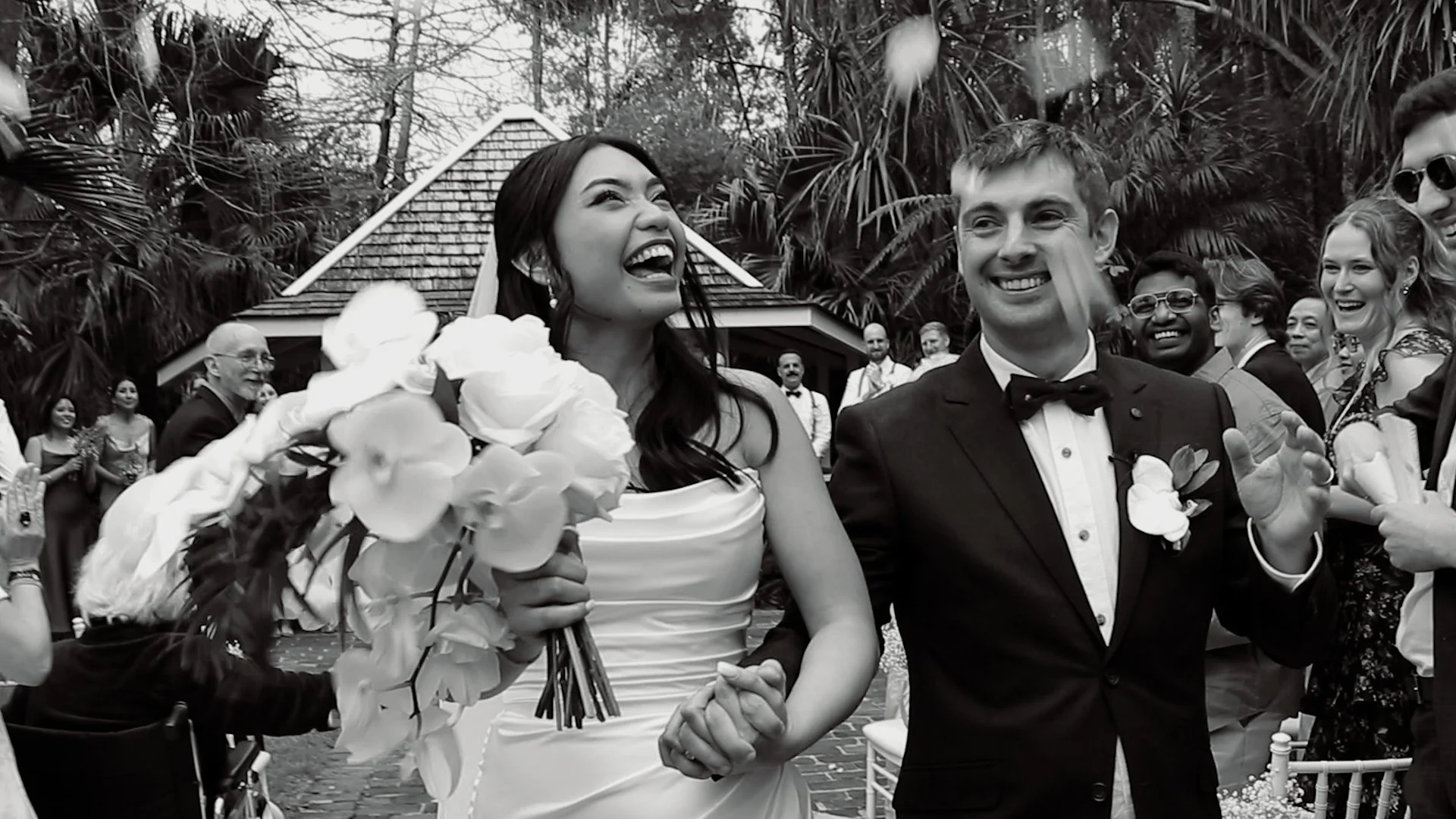 Black and white photo of a bride and groom smiling and holding hands at their wedding celebration surrounded by guests, palm trees, and a small chapel in the background.
