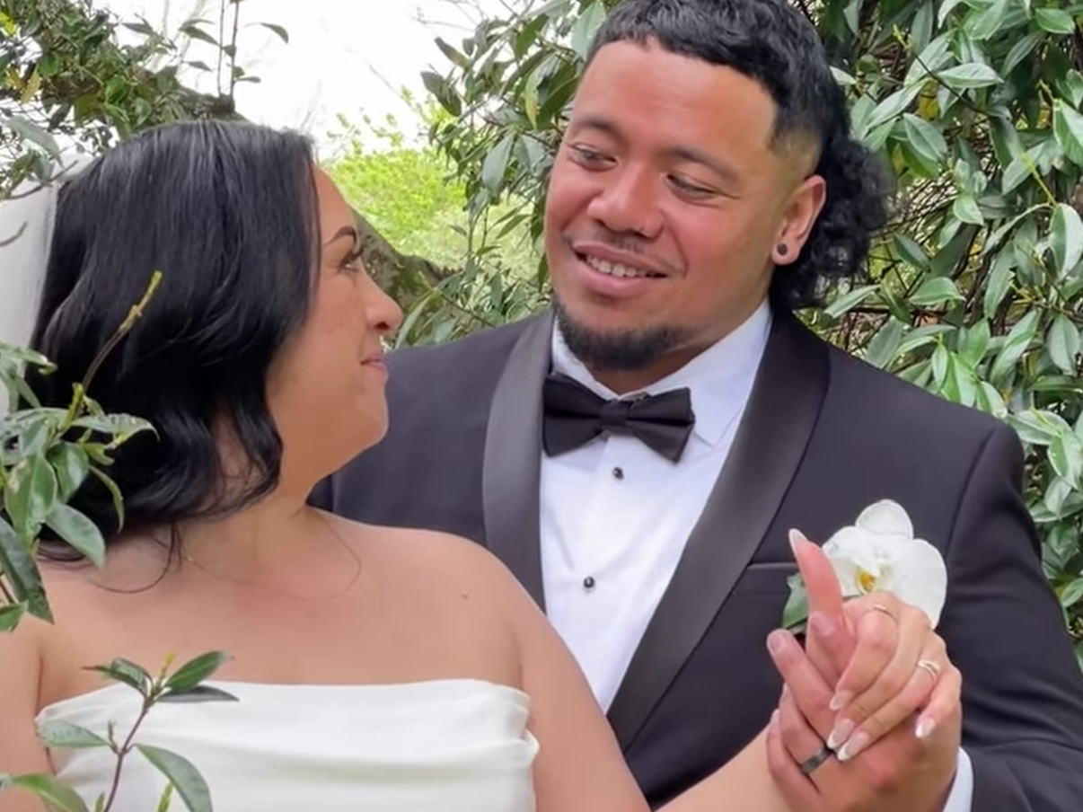 A bride and groom sharing a dance outdoors surrounded by greenery, with the groom holding the bride's hand and smiling.