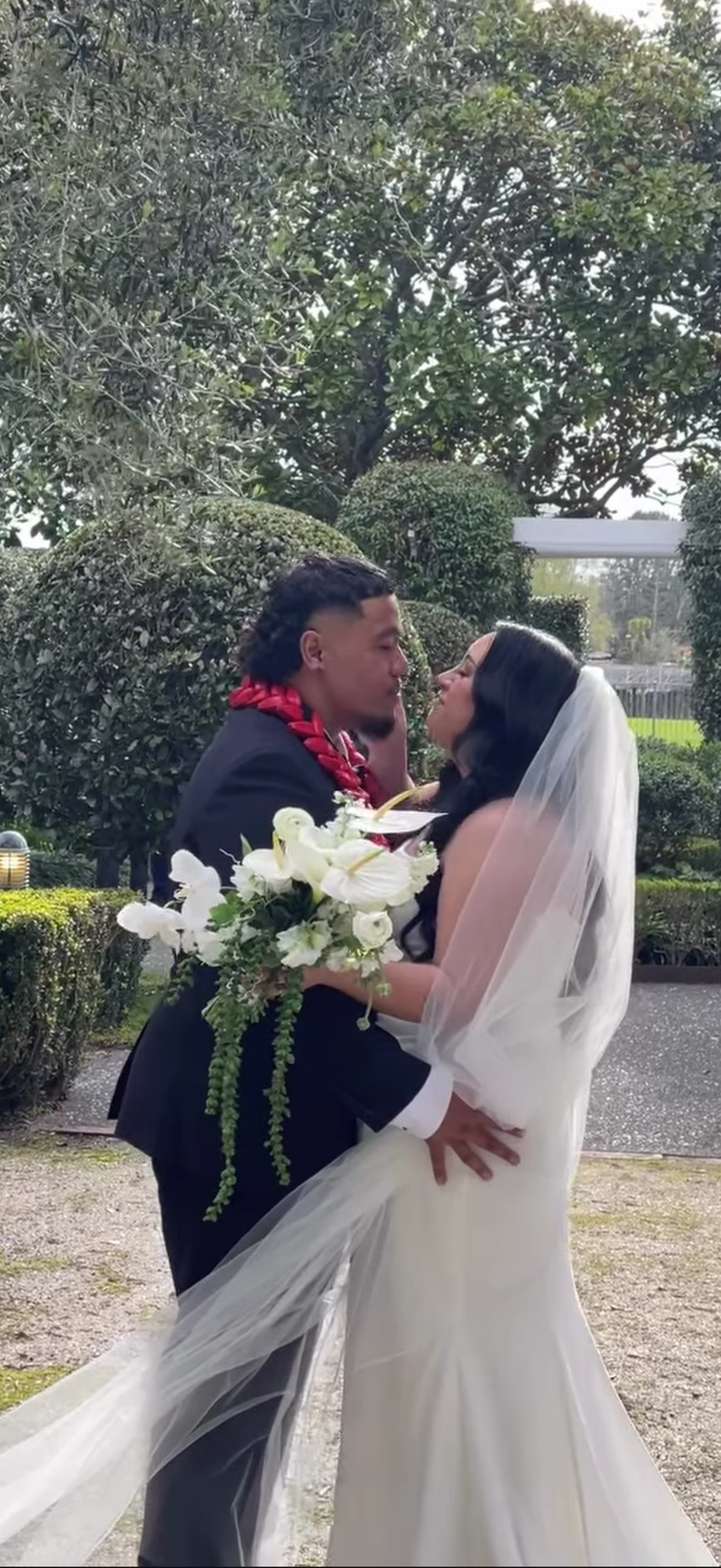 A couple dressed in wedding attire sharing a kiss outdoors, with the bride holding a bouquet of white flowers and wearing a white veil, and the groom wearing a black suit.