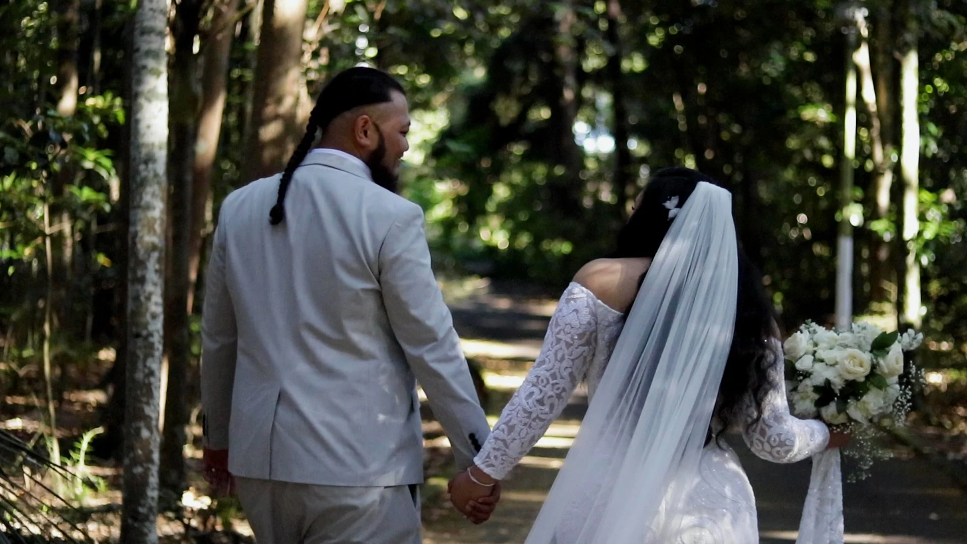 A newlywed couple holding hands and walking through a forested area. The bride is dressed in a white lace wedding gown with a long veil and holding a bouquet of white flowers. The groom is wearing a light-colored suit and has long braided hair.