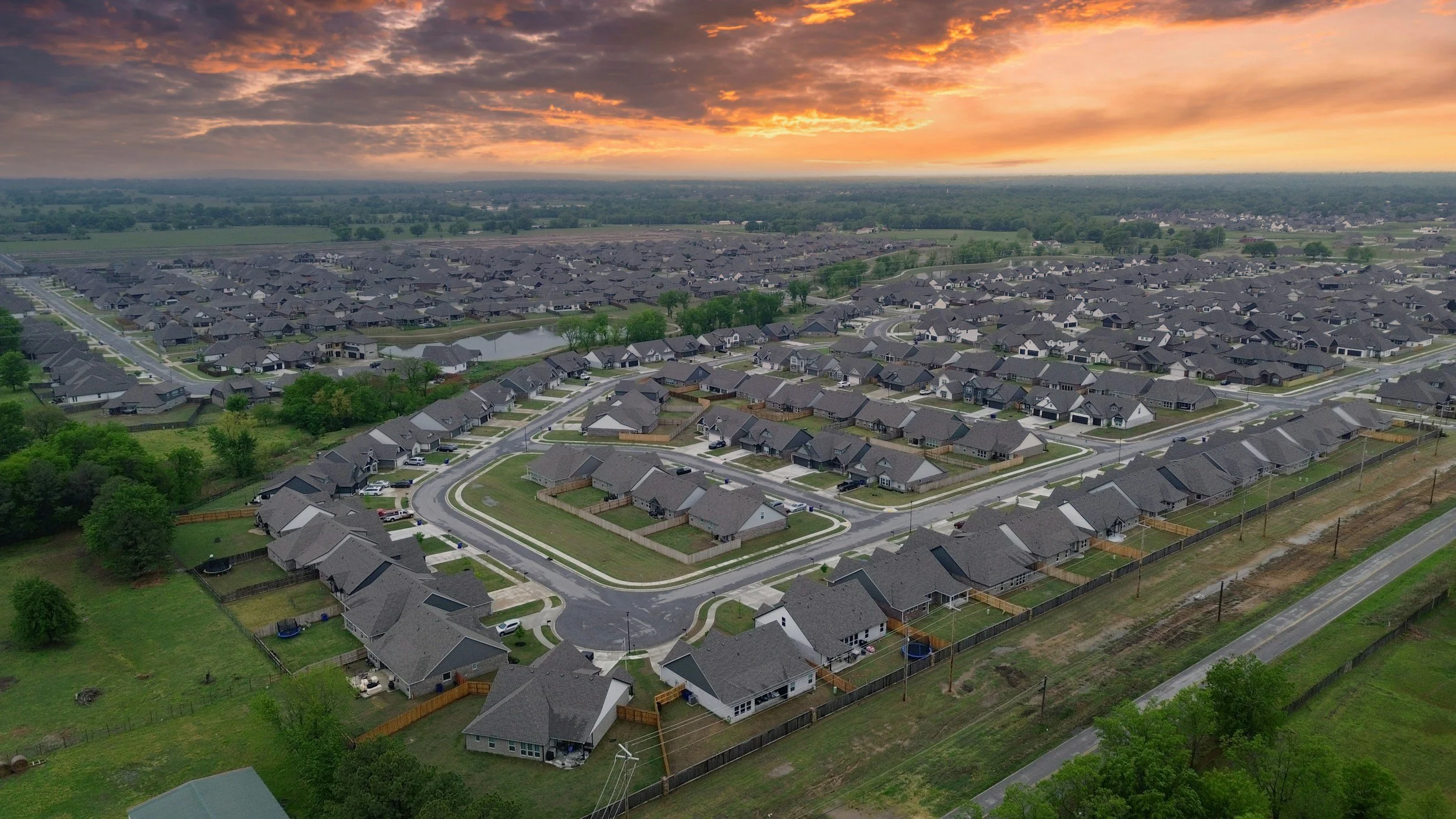 Aerial view of a suburban neighborhood at sunset with numerous houses, streets, green lawns, and trees; cloudy sky with orange and purple hues.