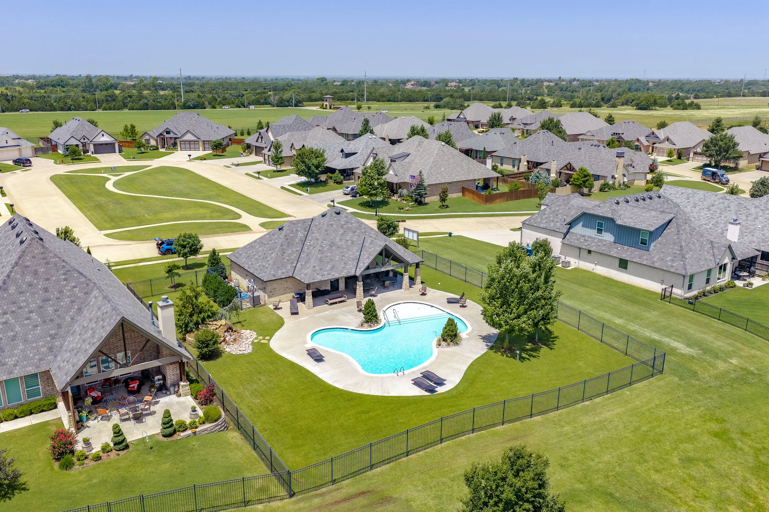 Aerial view of a suburban neighborhood with houses, green lawns, a swimming pool, and a fenced backyard on a sunny day.