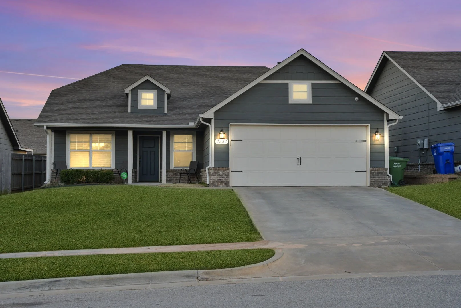Gray single-family house with a front yard, driveway, and attached garage, illuminated by exterior lights at dusk, with a purple and pink sunset sky in the background.