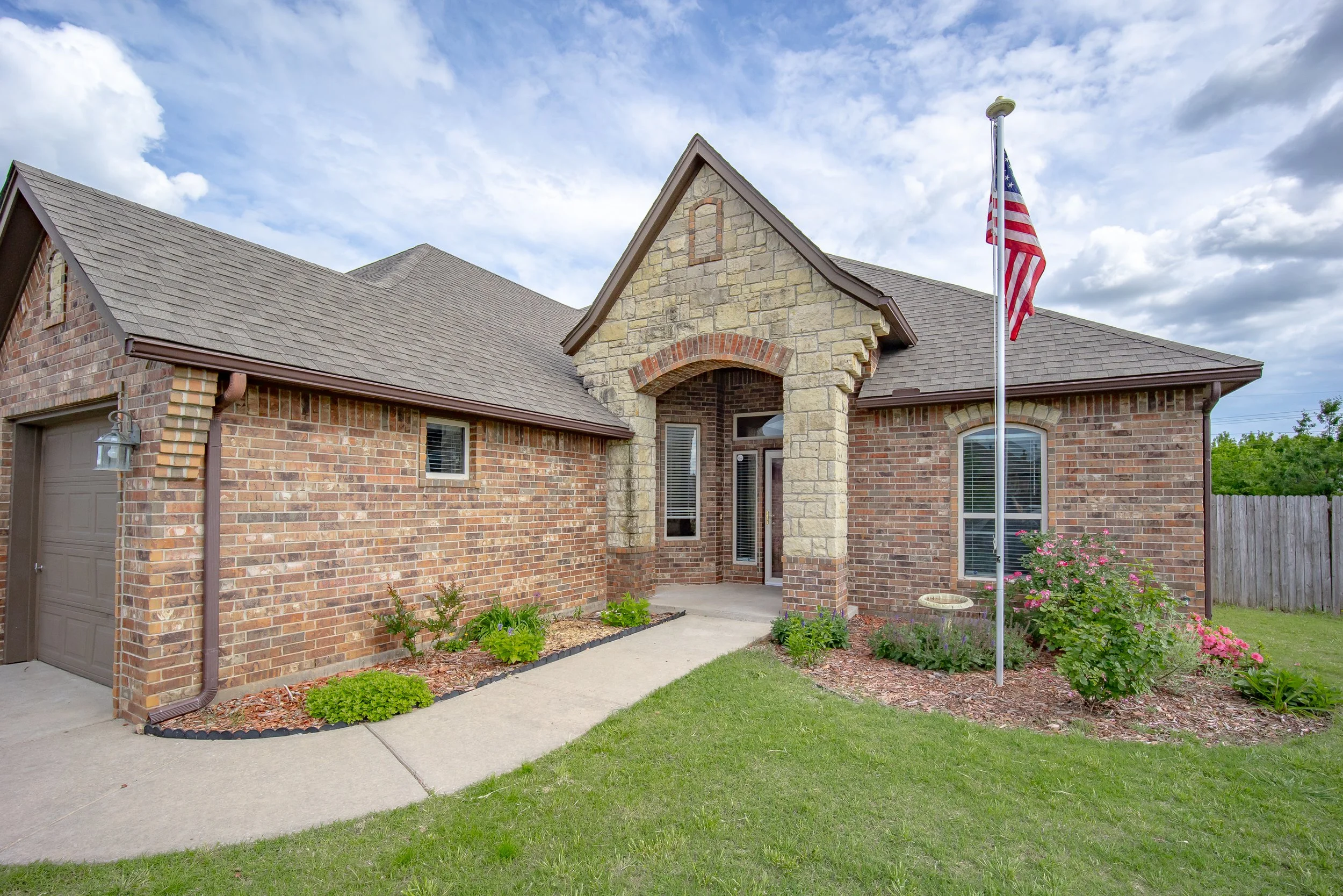 Front view of a brick house with a stone entrance, American flag on a flagpole, garden beds with flowers, a concrete sidewalk, and a lawn on a partly cloudy day.