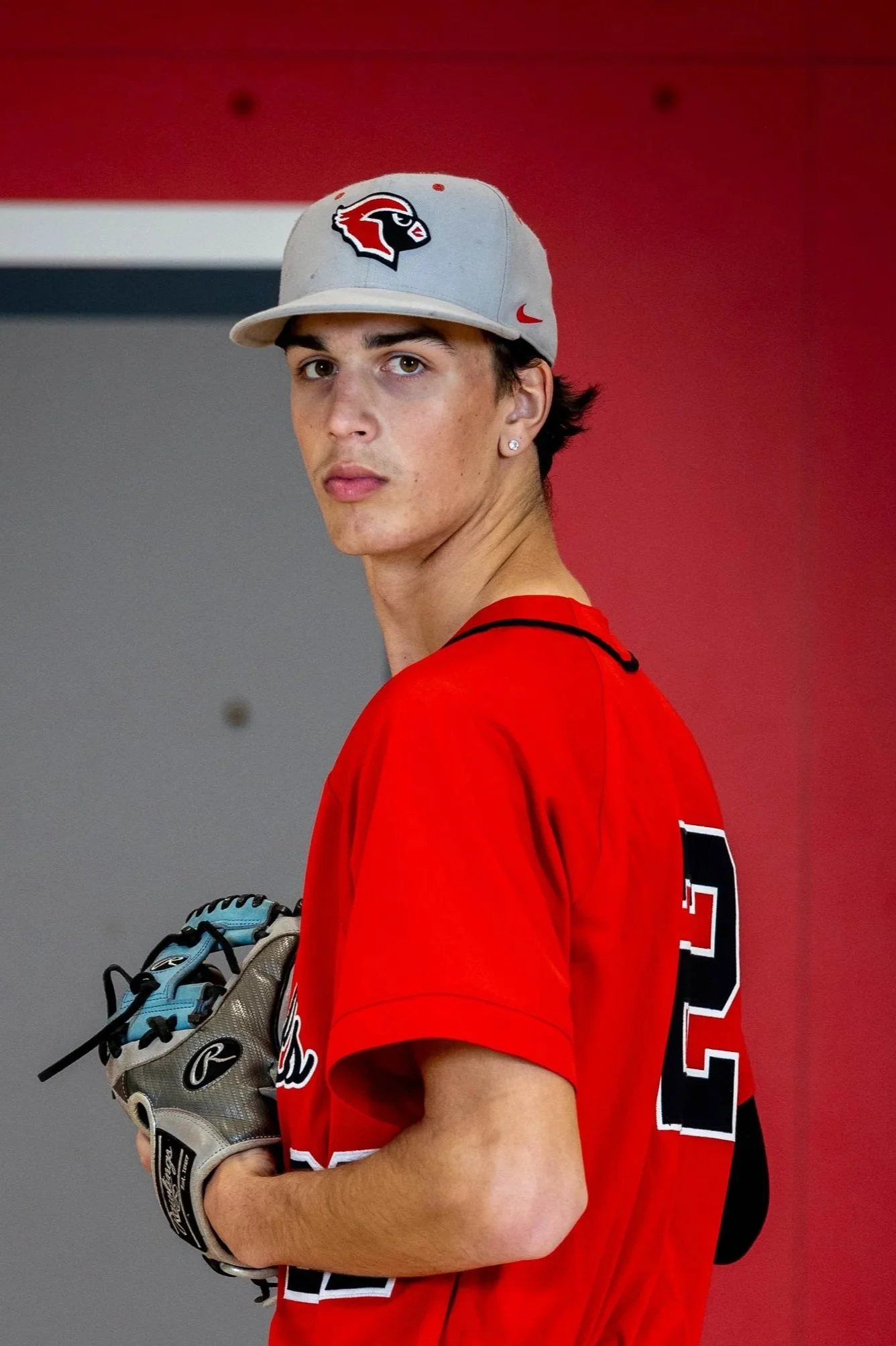 Young male baseball player in a black and red uniform holding a baseball and a glove, wearing a cap with a red and white logo, against a red background.