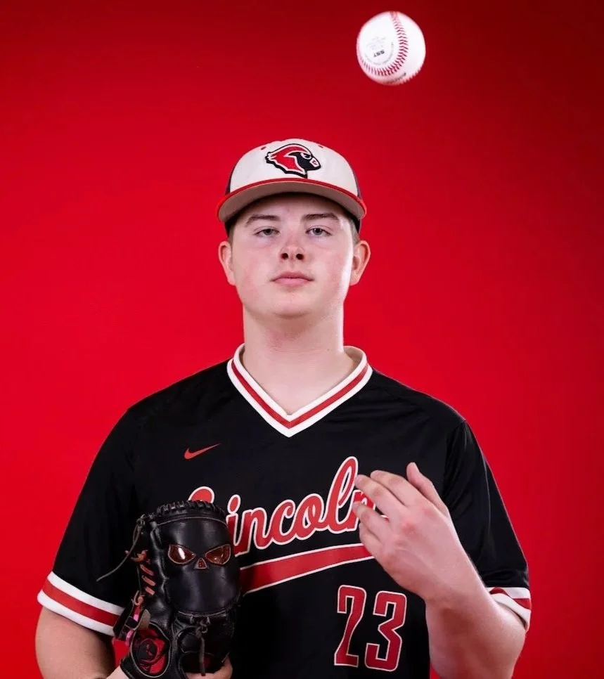 Young male baseball player in black and red uniform, holding a black glove, wearing a red and white cap, standing against a red background.