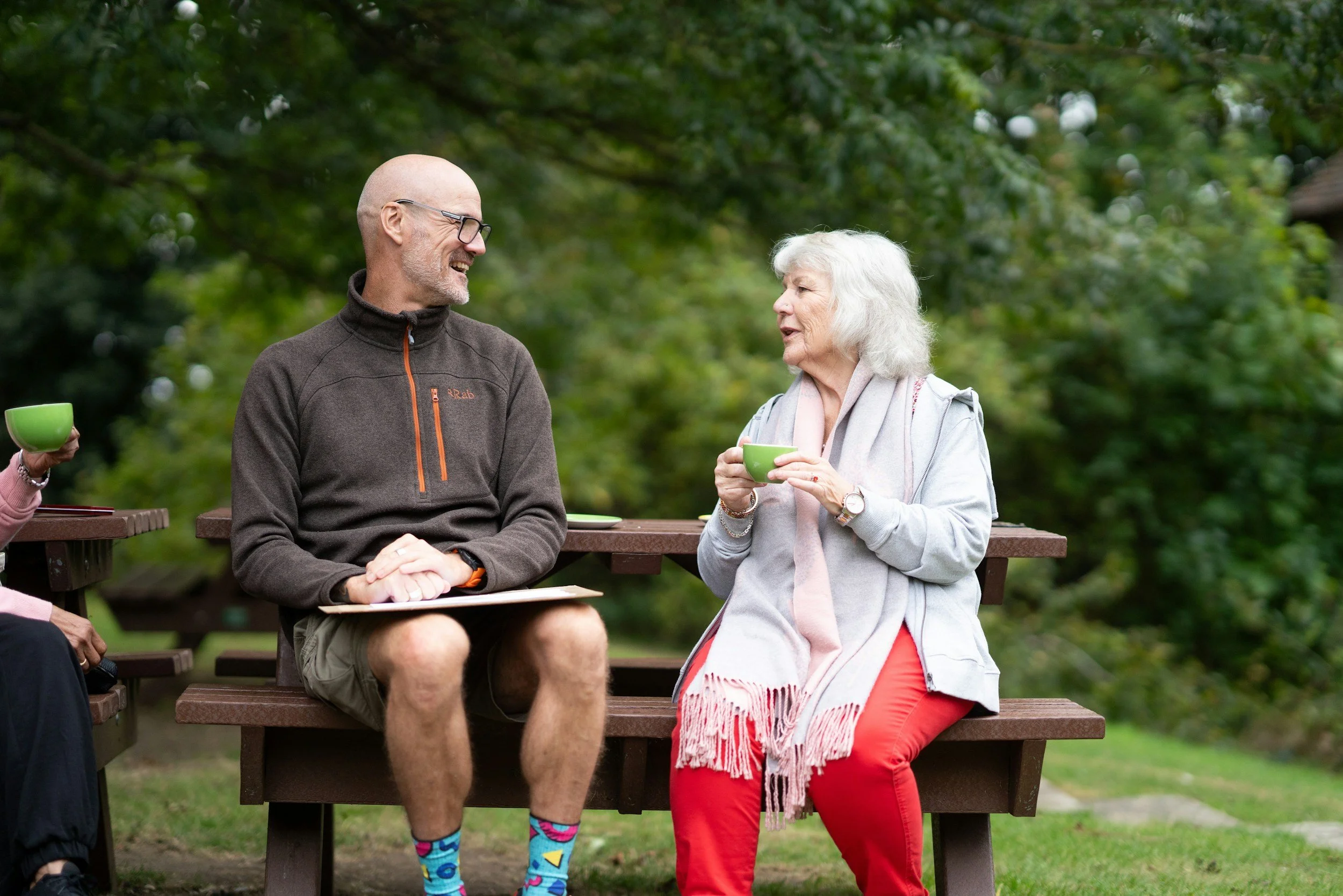 Two elderly women and a middle-aged man sitting on benches outdoors, engaged in conversation, with one woman holding a green cup, surrounded by greenery.