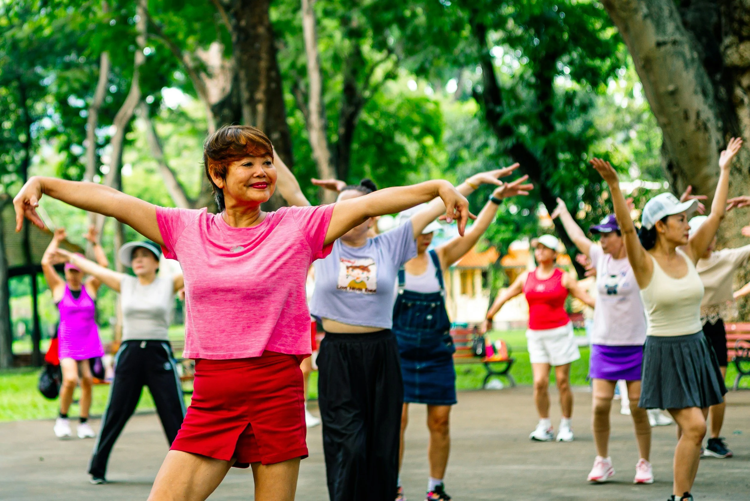 A group of women participating in outdoor exercise or dance class in a park, stretching with arms raised, surrounded by lush green trees.