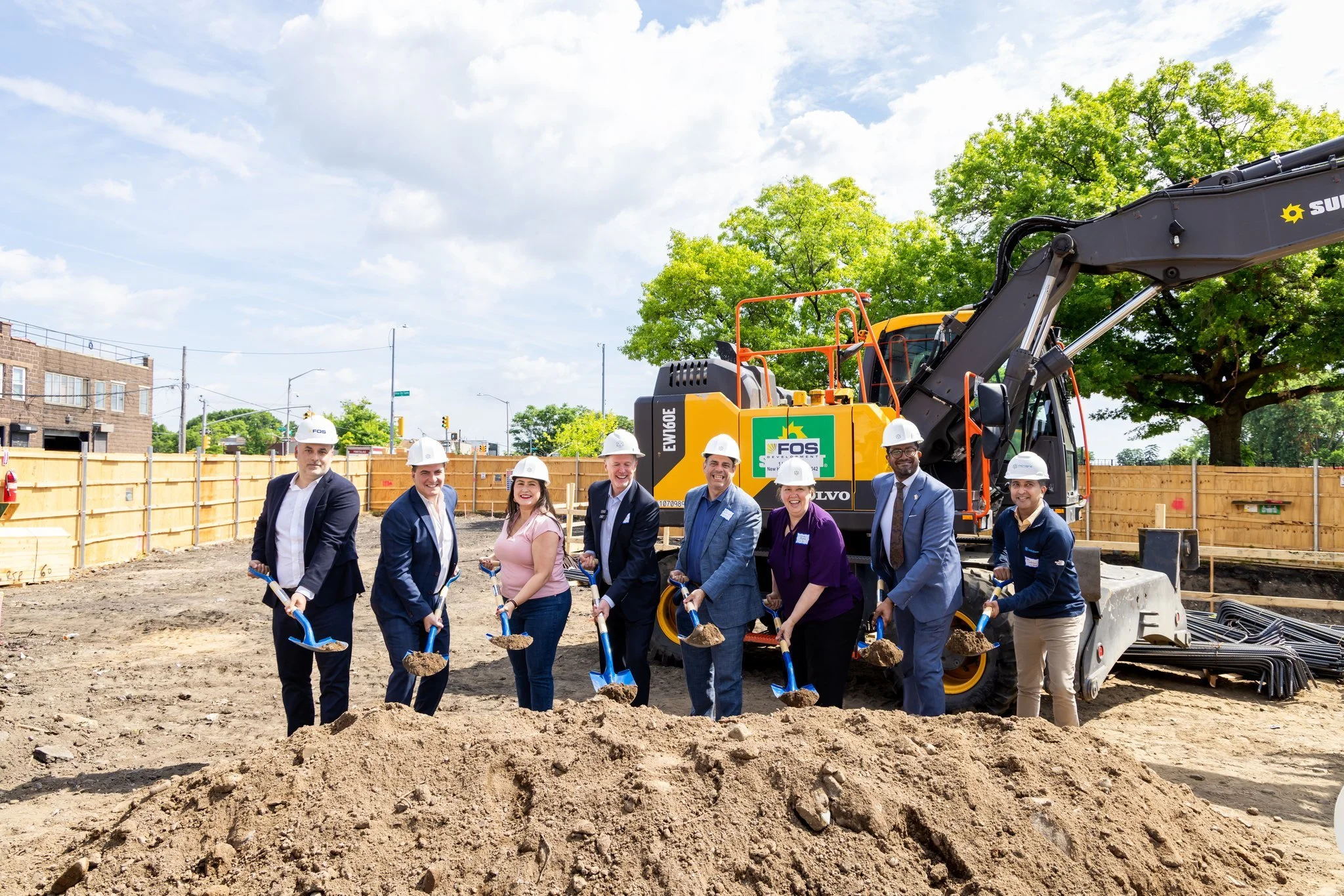 A group of people in business attire and construction helmets standing on a construction site with shovels, smiling. Behind them is heavy construction equipment, a wooden fence, trees, and a partly cloudy sky.