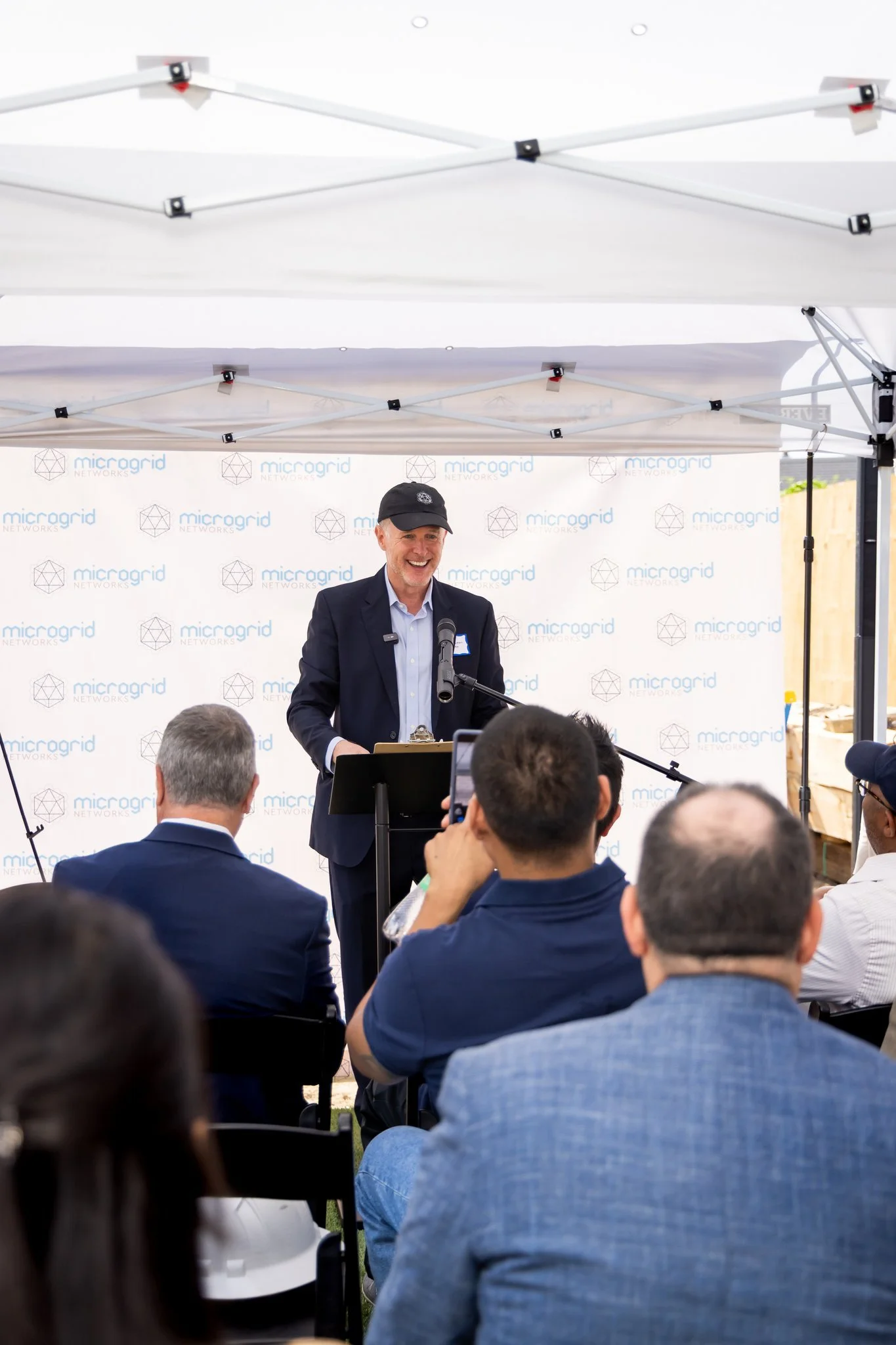 A man in a dark suit and cap speaking at a podium during an outdoor event. The backdrop displays the 'microgrid NETWORKS' logo repeatedly. Audience members are seated, some taking photos or recording.