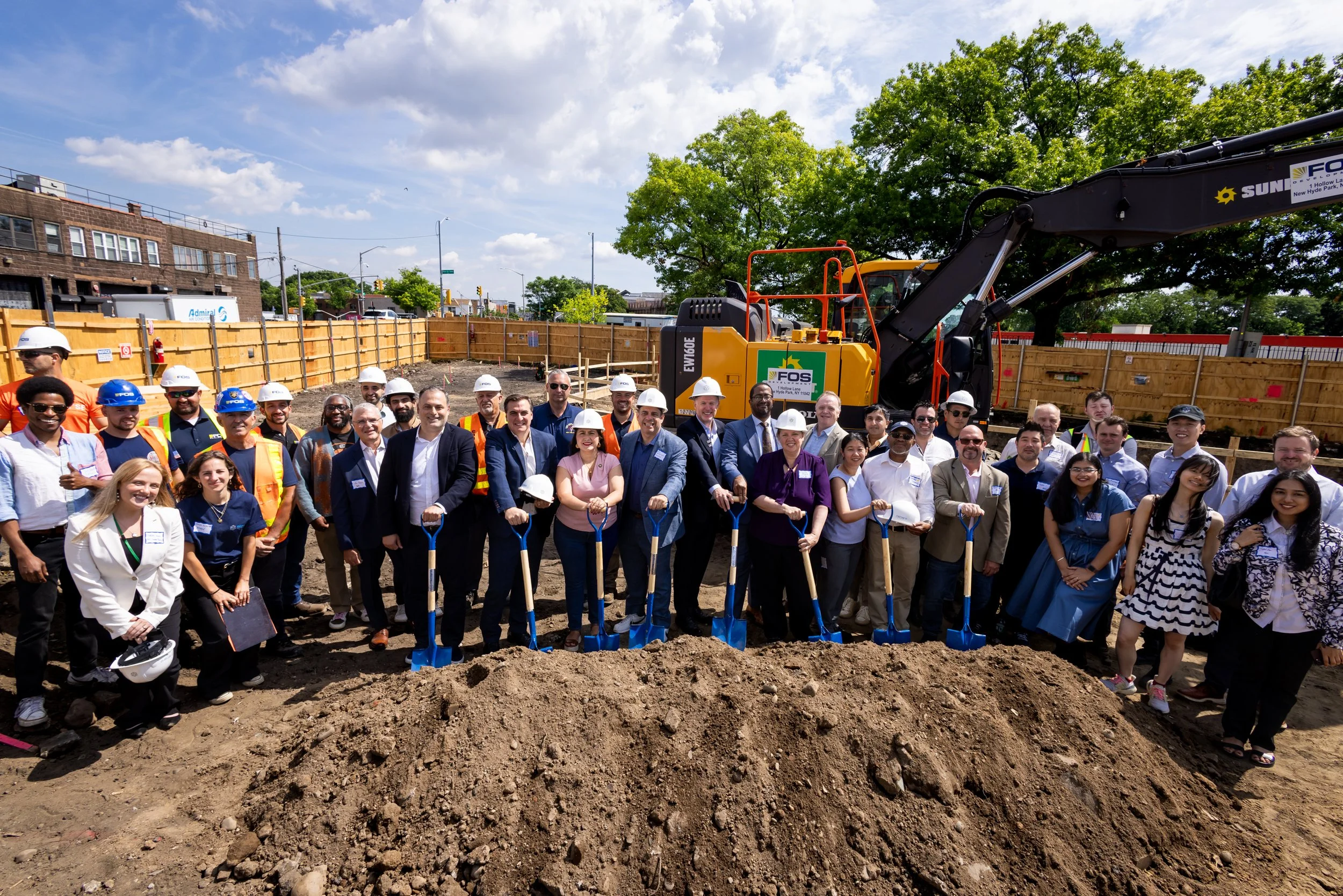 A large group of people, including construction workers wearing safety helmets, gathered together at a construction site during daytime for a groundbreaking ceremony. There are shovels in front of them, and a construction vehicle is visible in the background. The sky is partly cloudy, and there are trees and buildings behind the site.