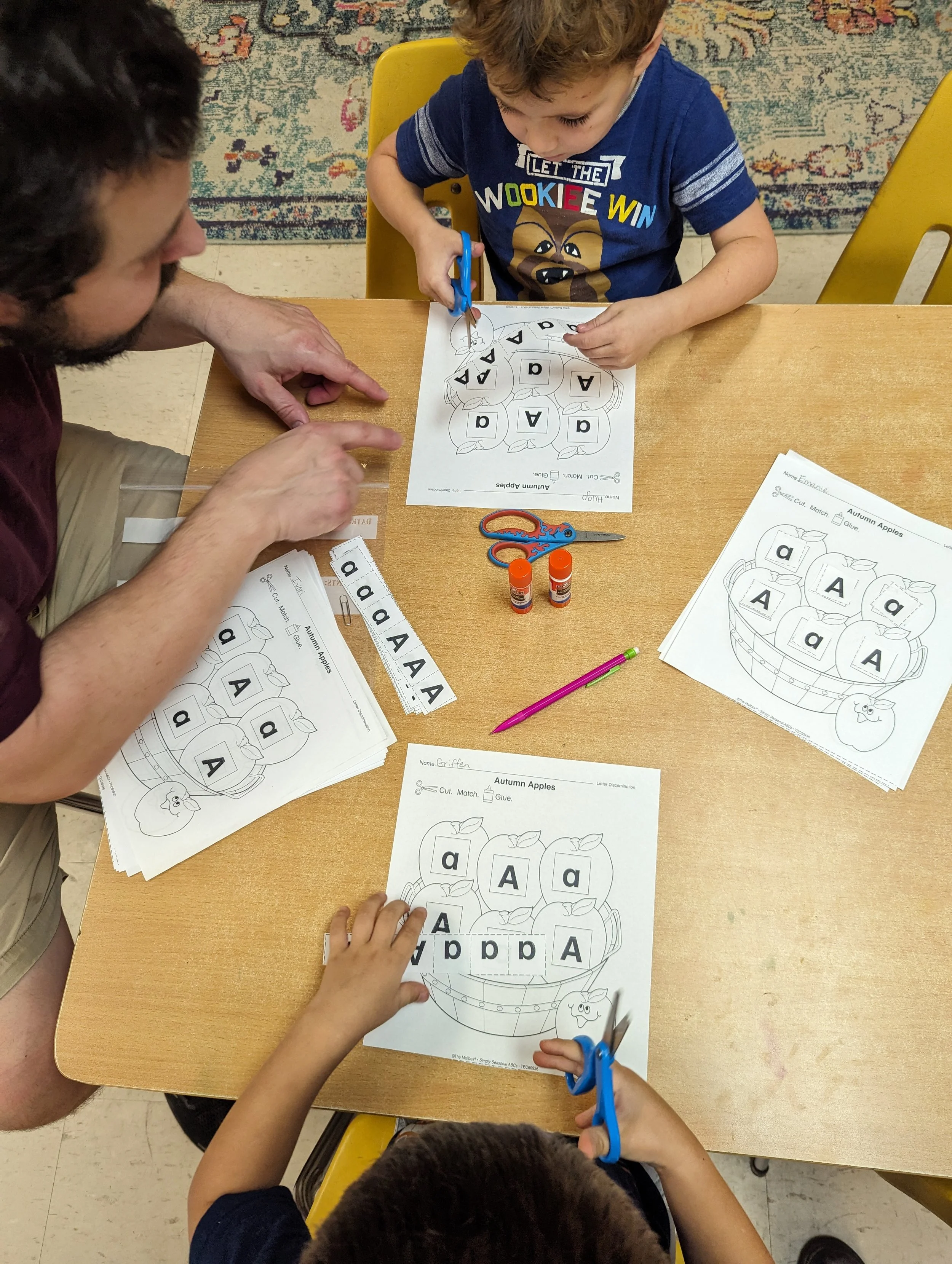 Children and an adult cutting and gluing letter cutouts on a themed worksheet.