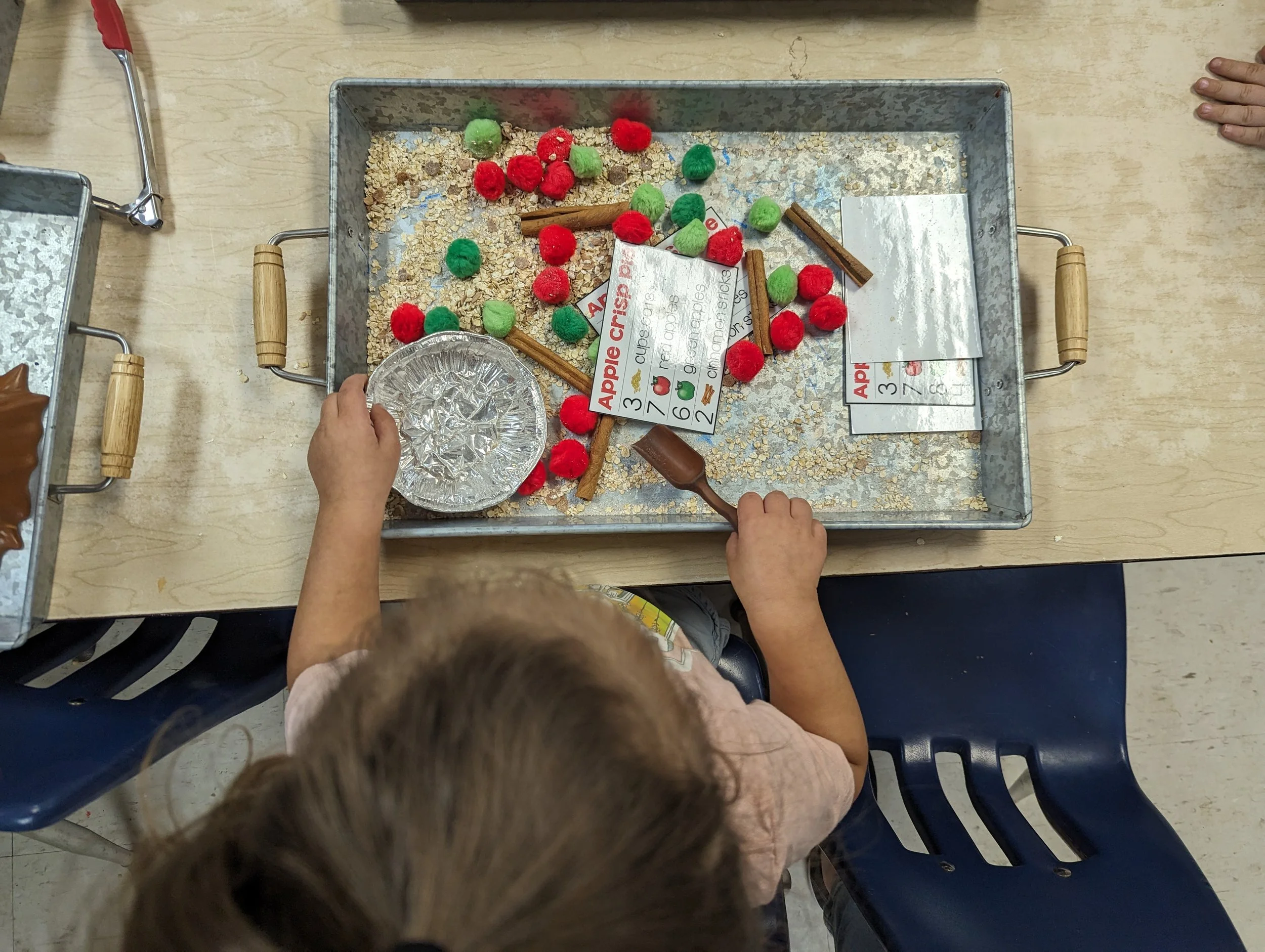 Child playing with a sensory bin filled with oats, red and green pom-poms, cinnamon sticks, and a small aluminum pie plate, on a wooden table.