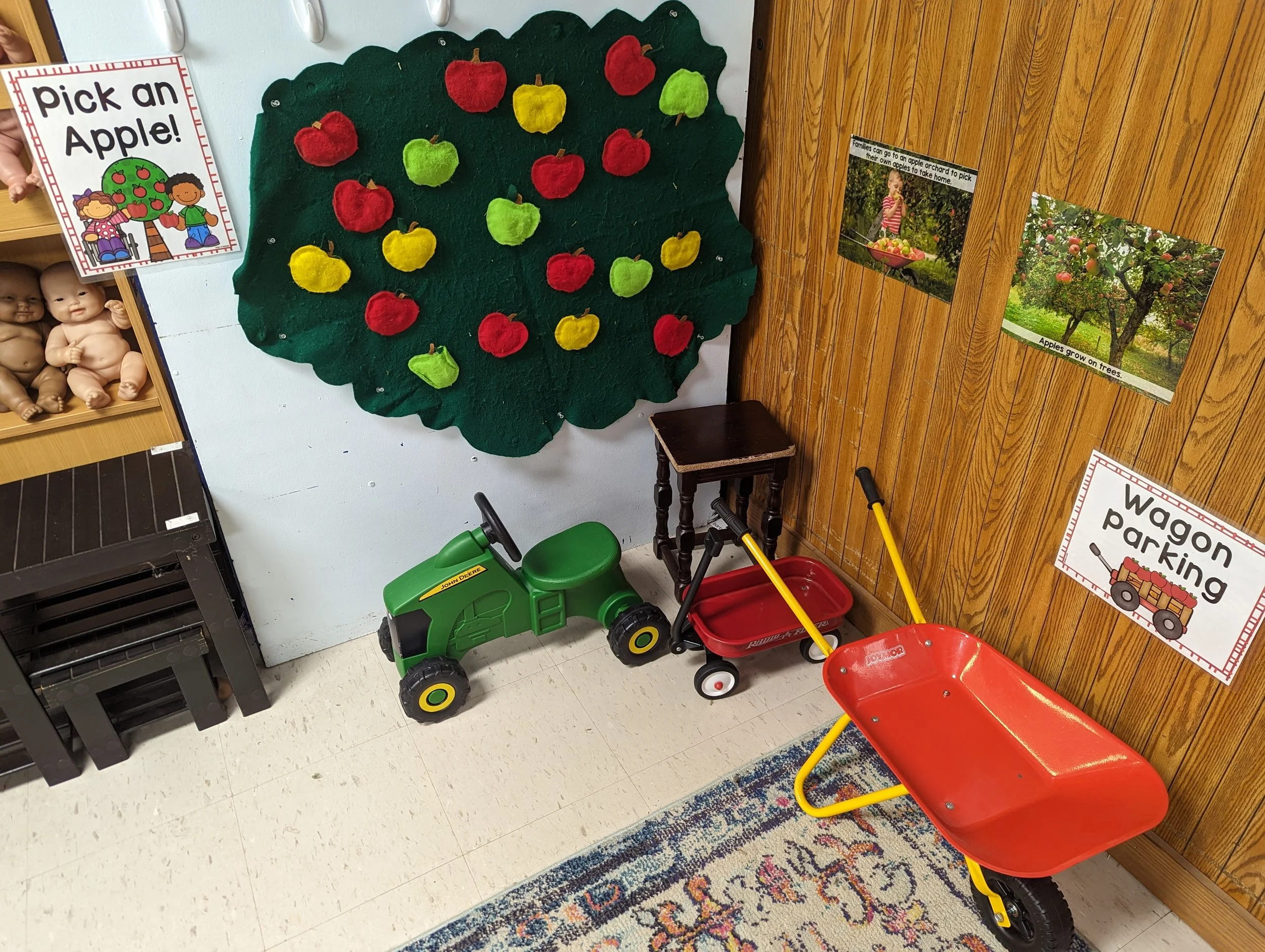 Toy indoor play area with a felt apple tree decoration, a green toy tractor, a red wagon, and a red cart.