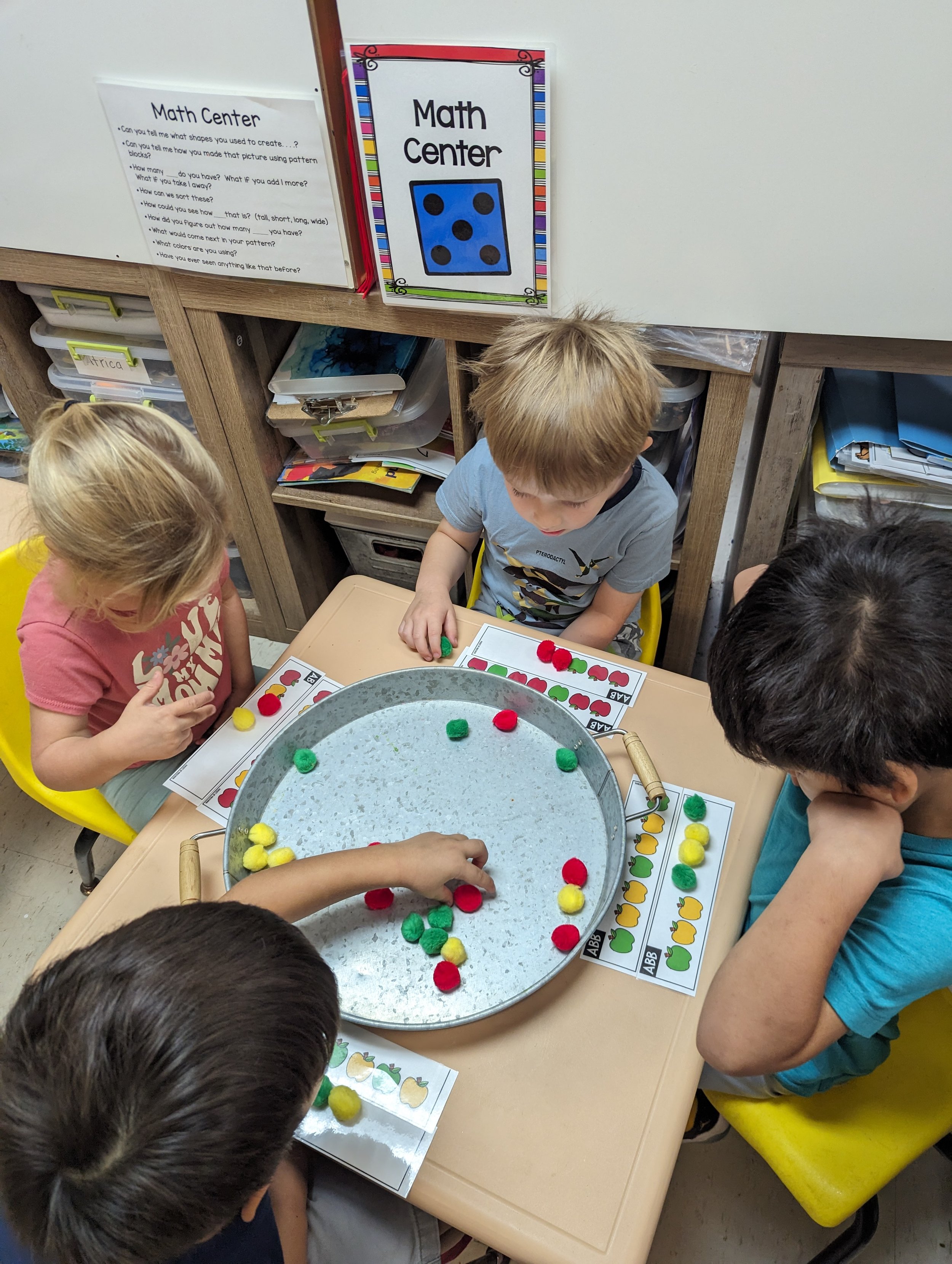 Children playing a game with colored pom-poms and printable sheets at a classroom table.