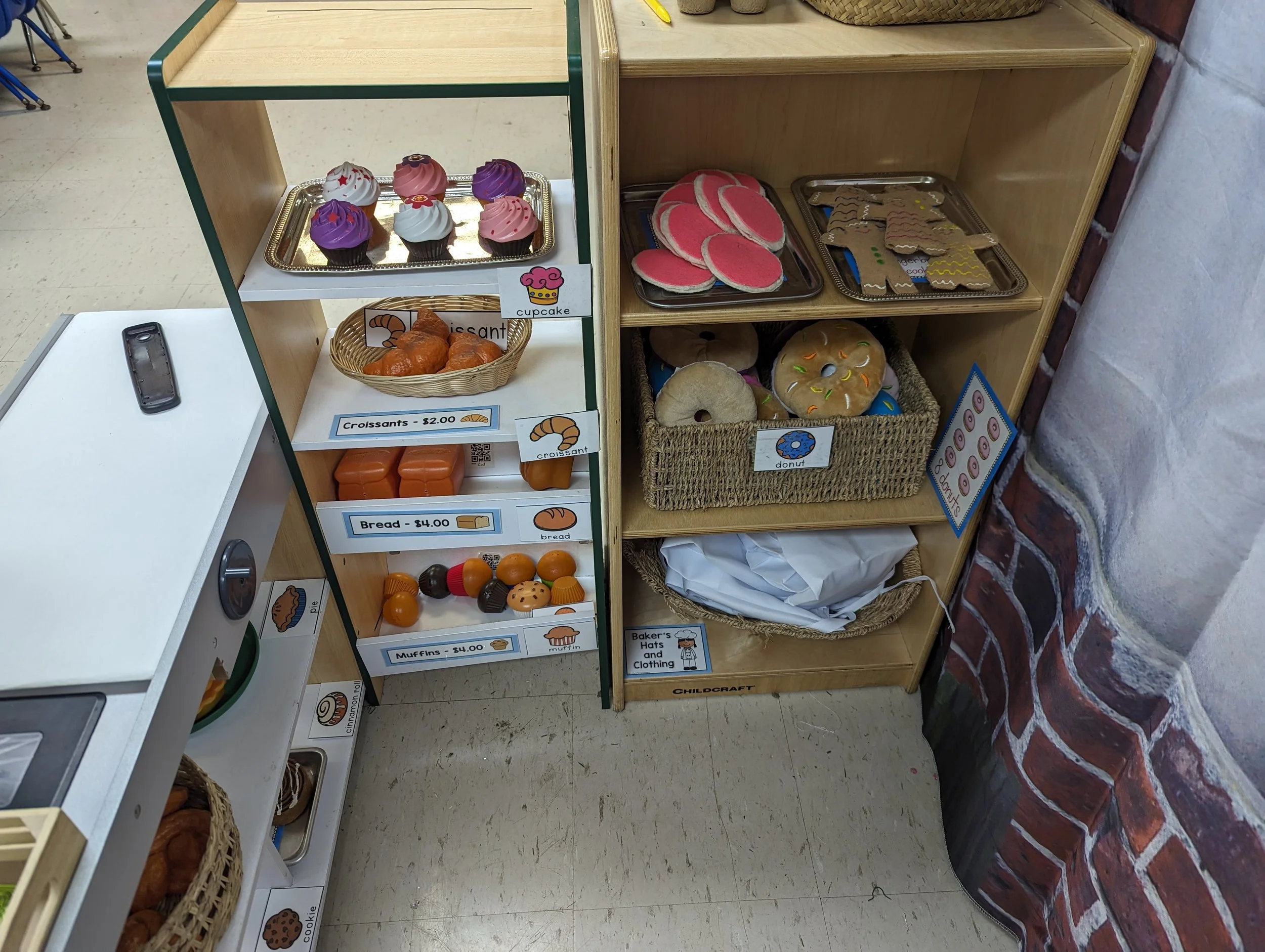 A display of pretend baked goods on a wooden and metal shelving unit and table, including cupcakes, croissants, bread, muffins, and donuts.