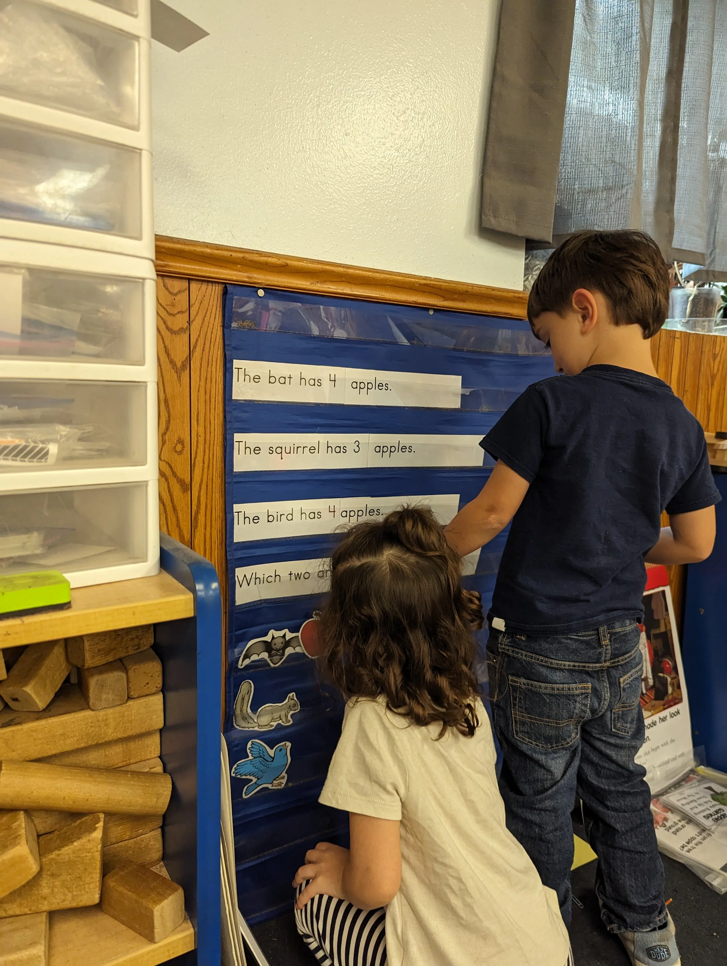 Two children standing and kneeling in front of a blue pocket chart.