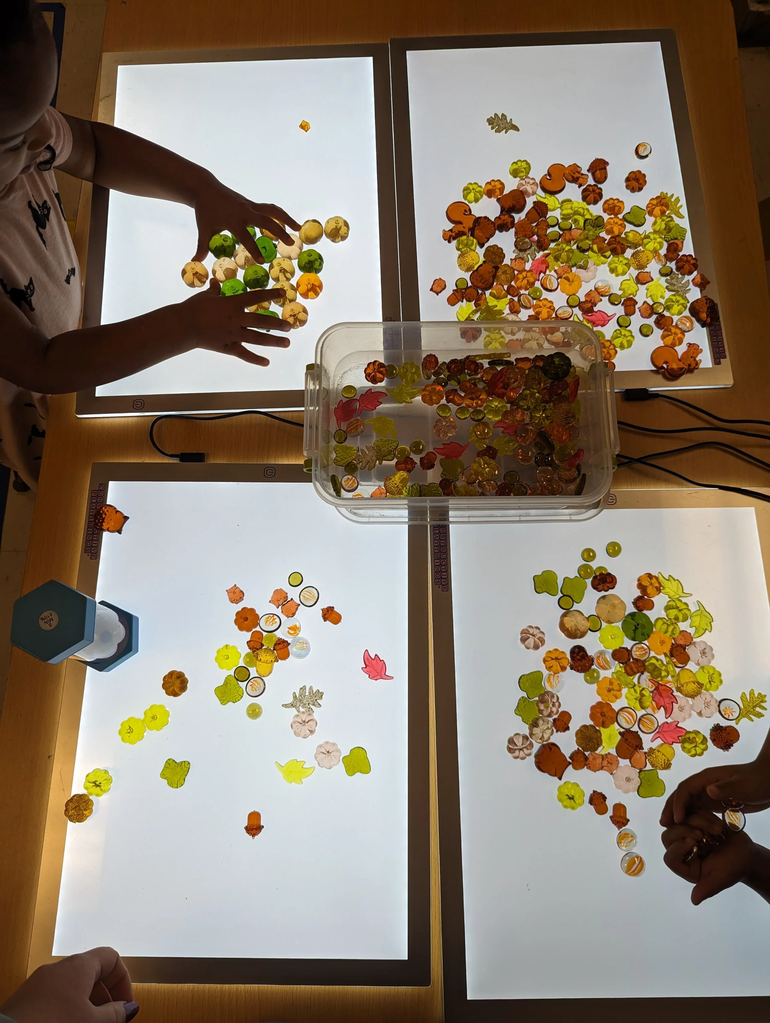 Children sorting and playing with colorful leaf and acorn-shaped cutouts on lightbox tables, with a container of additional cutouts in the center.