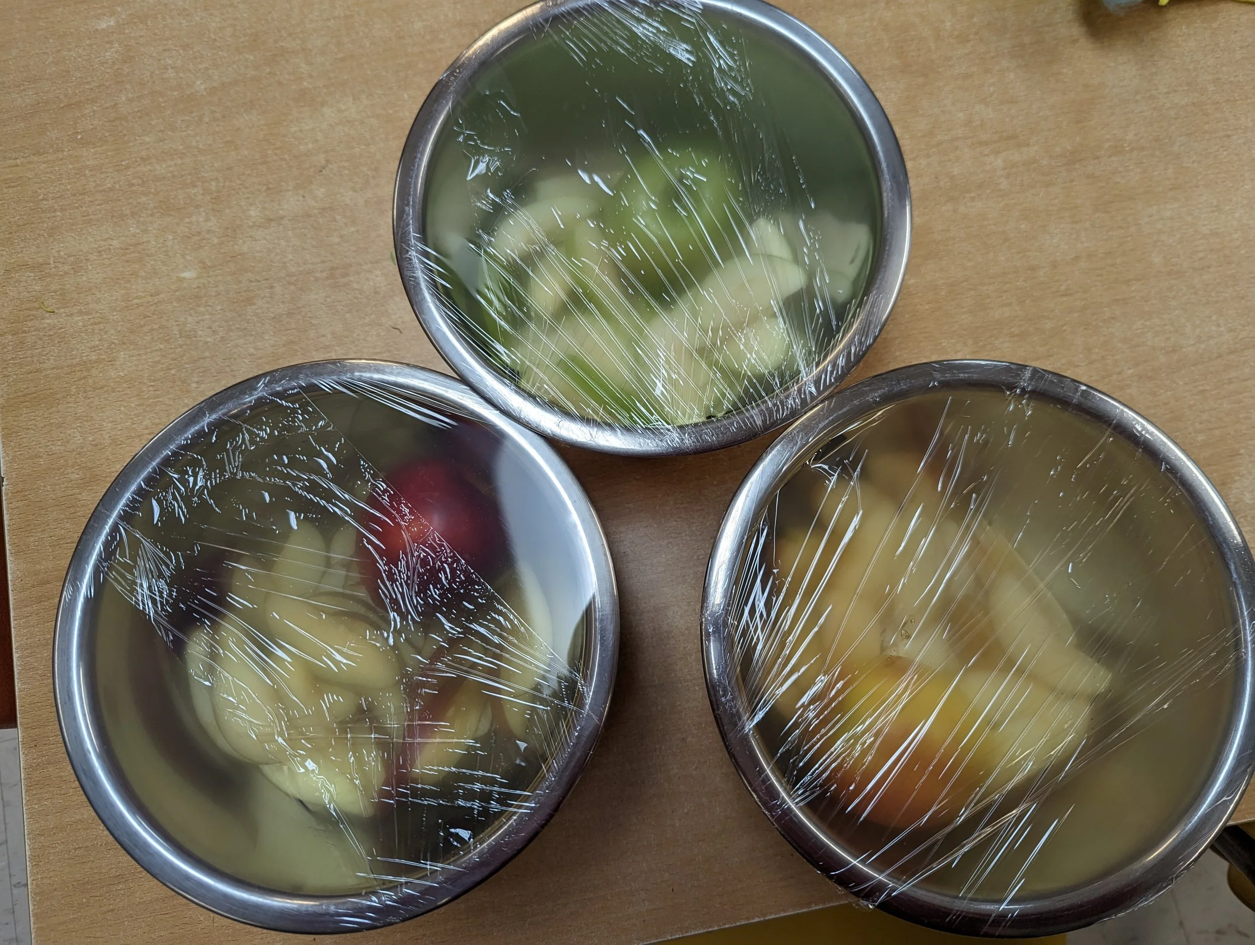 Three stainless steel bowls contain cut fruits, covered with plastic wrap.