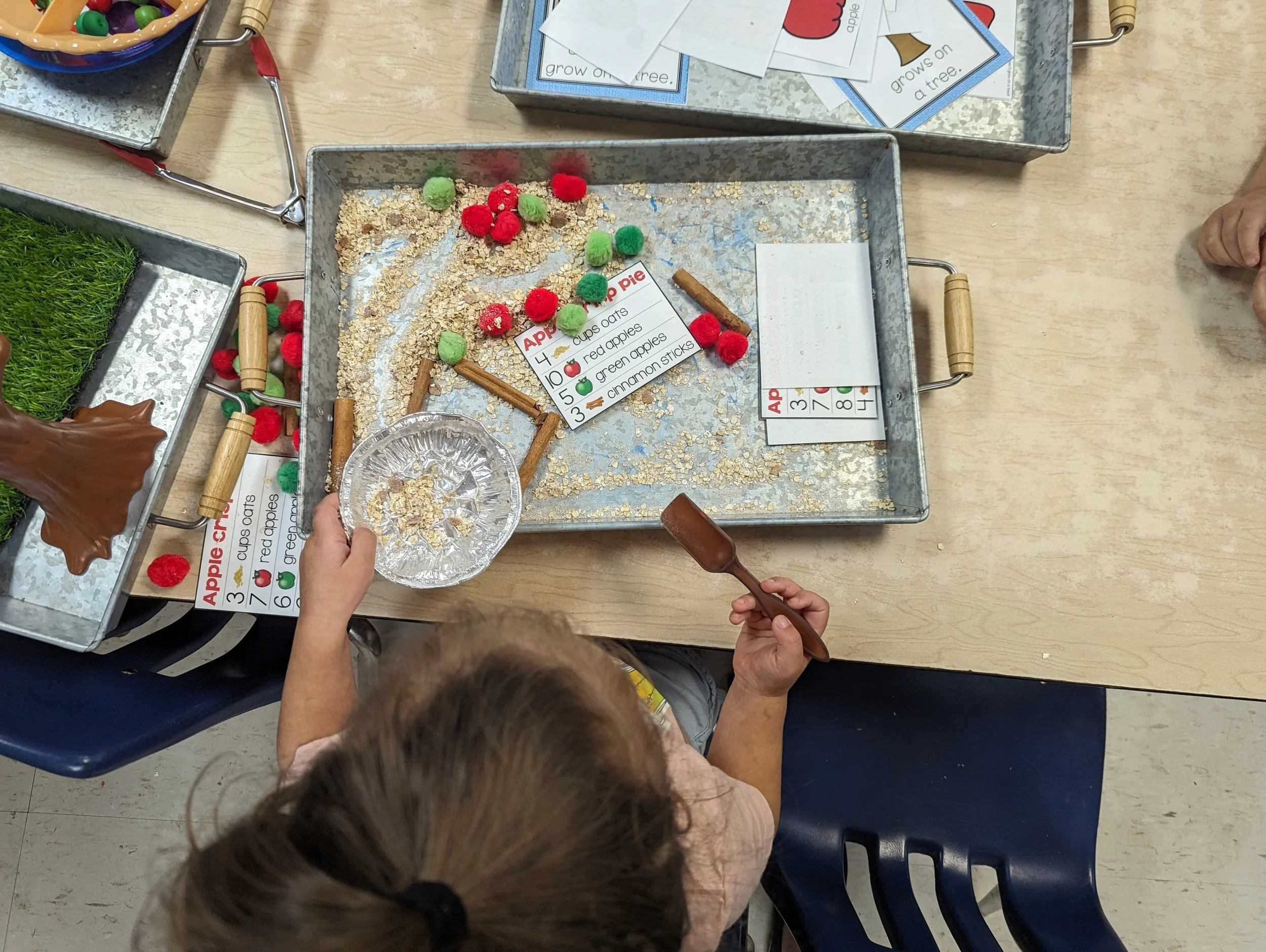 Child using a small spoon to scoop oats and a pie crust from a tray, with colorful pom-poms and cinnamon sticks on the tray, for a pretend apple pie activity.