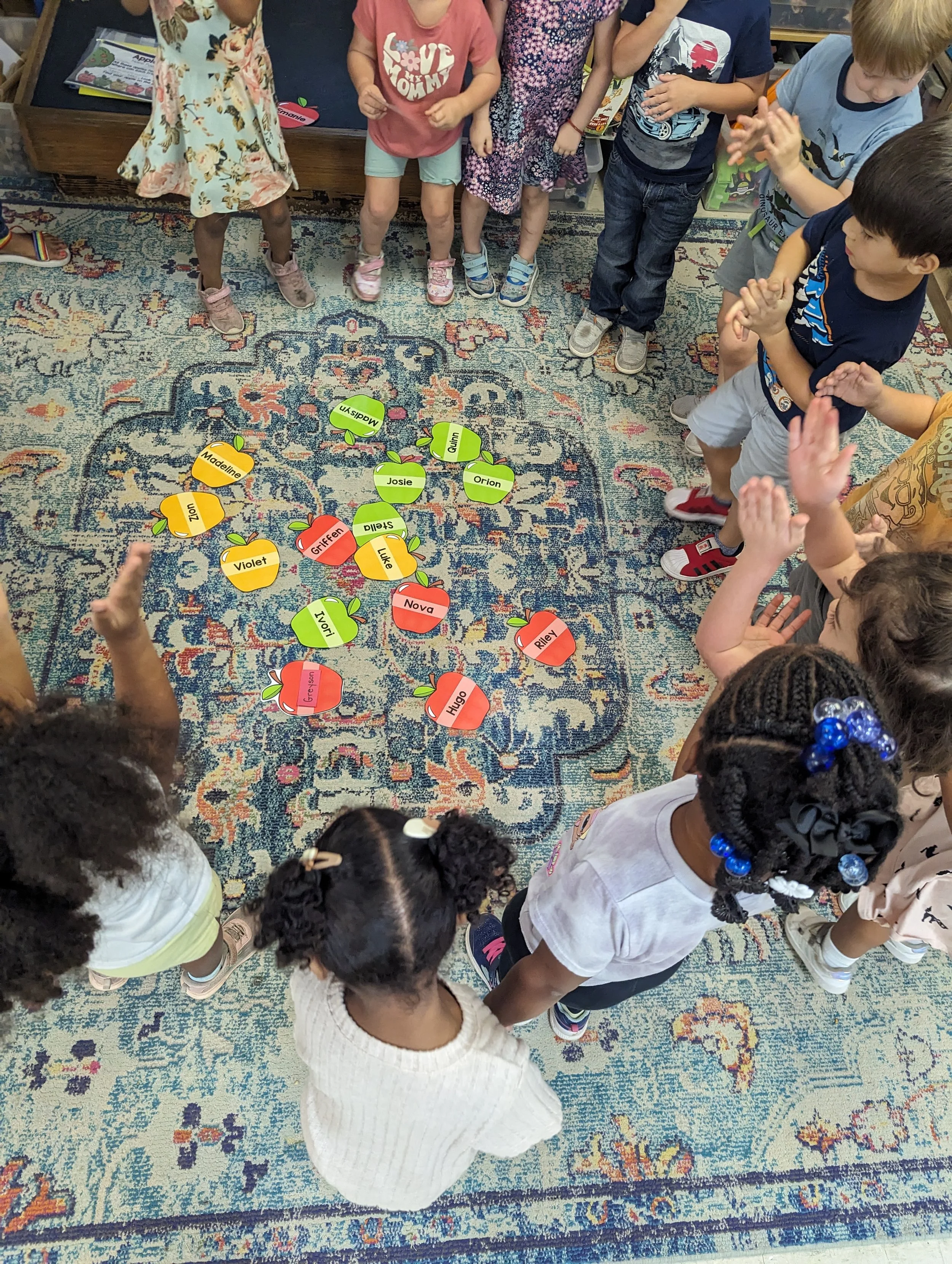 Children gathered around a colorful carpet with paper apple cutouts on the floor, engaged in a group activity.