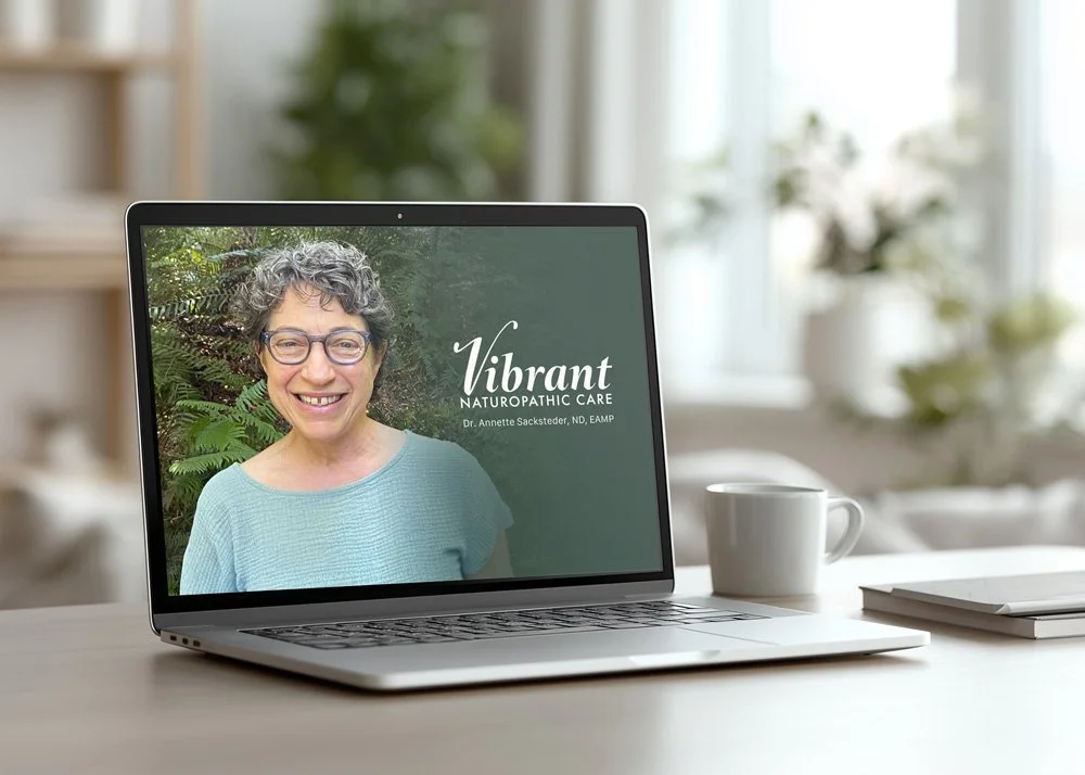 Laptop on a desk displaying a smiling woman with curly gray hair and glasses, with green foliage behind her. The screen text reads 'Vibrant Naturopathic Care, Dr. Annette Sacksteder, ND, EAMP.'