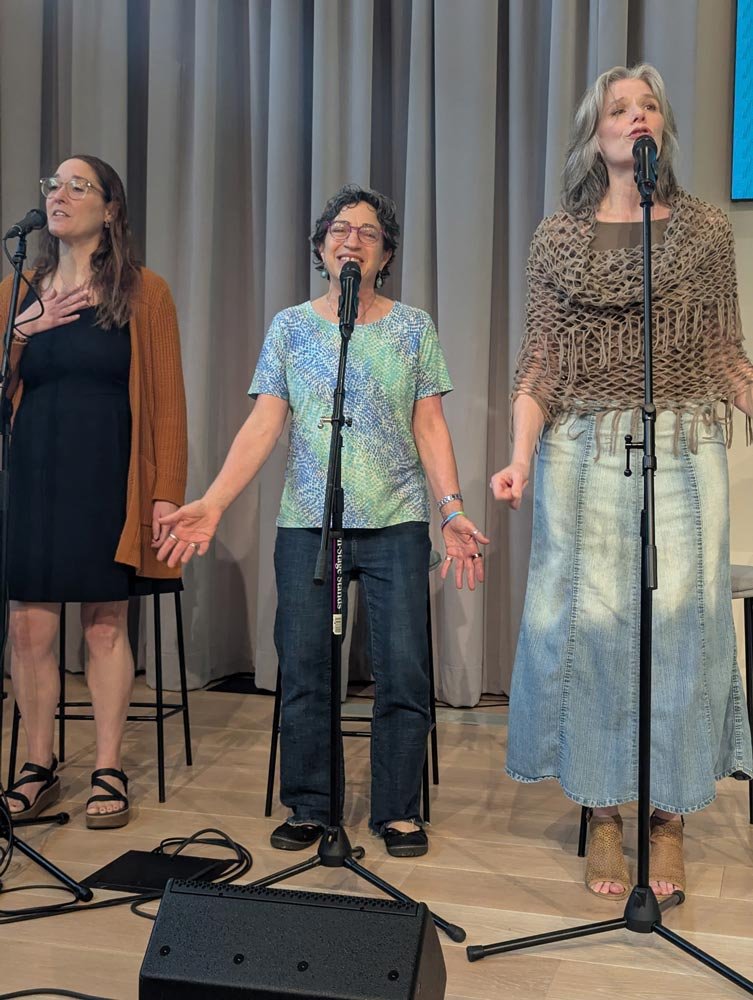 Three women singing on a stage with microphones and a gray curtain background.