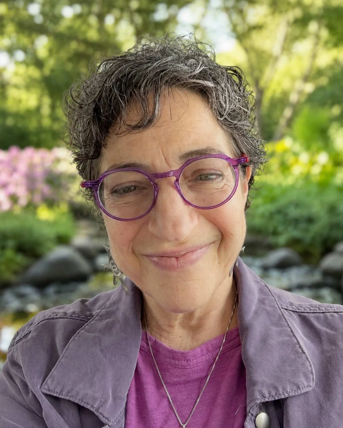Close-up of a smiling woman with short, curly gray hair and purple glasses, outdoors with a blurred background of green trees and rocks.
