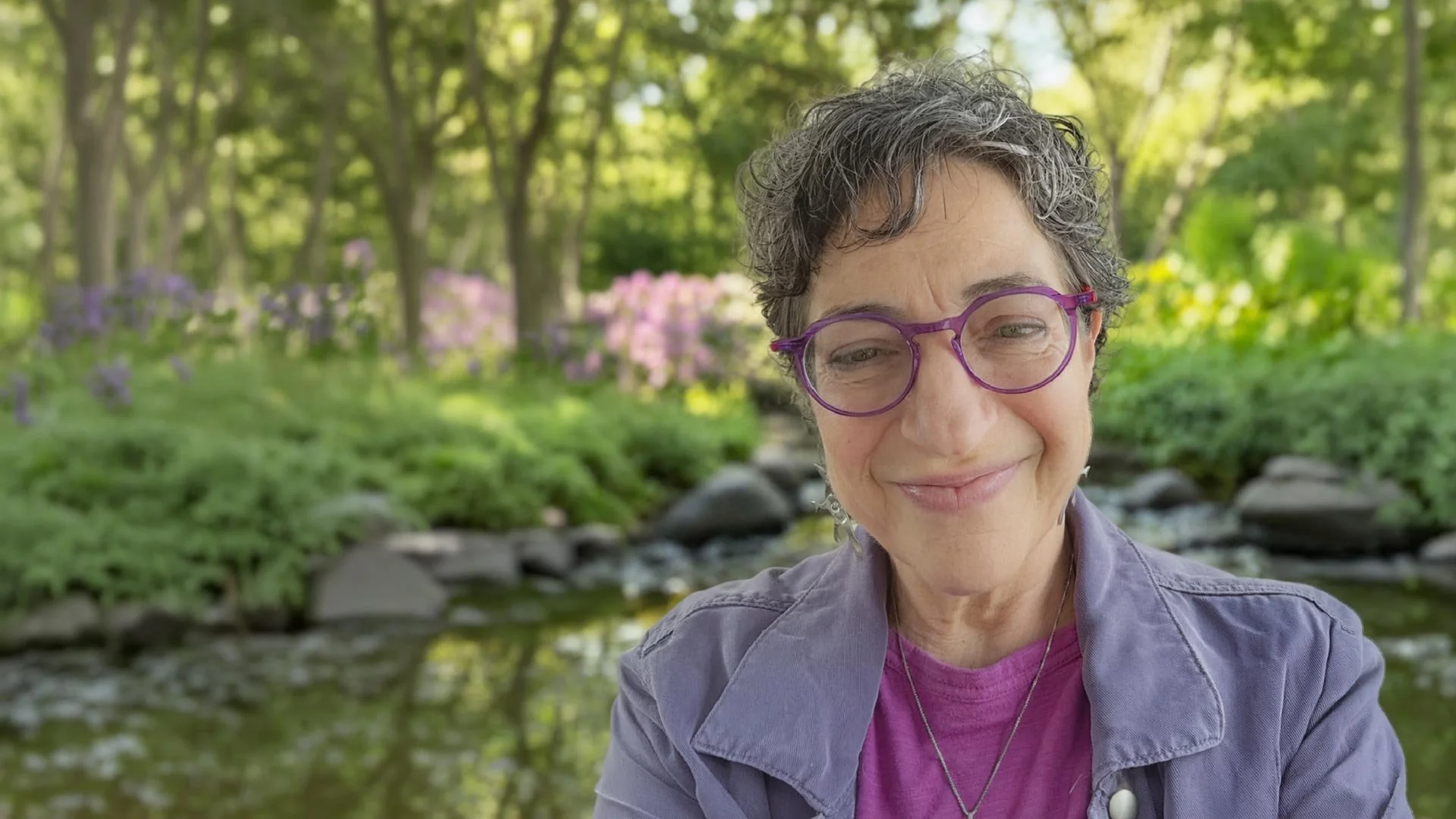 A middle-aged woman with short curly gray hair and purple glasses smiling outdoors near a stream, with trees and pink flowers in the background.